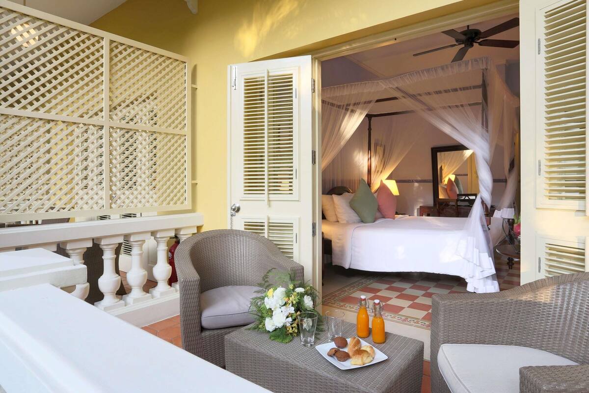 Ein Hotelzimmer mit Bett, einem Korbsessel, einem Tisch mit Frühstück und einem Balkon mit Gitterverkleidung.