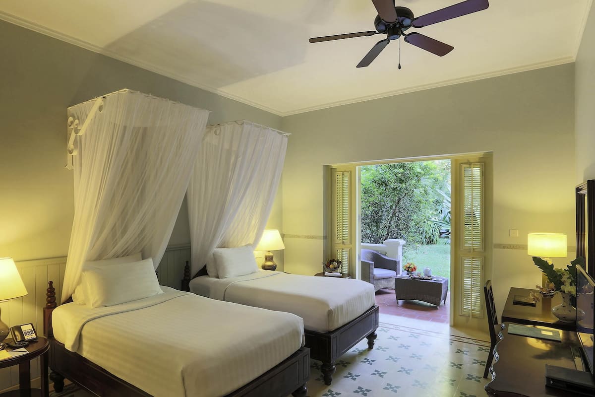 A bedroom with two beds, a ceiling fan, a nightstand with a lamp, and a view of the garden.
