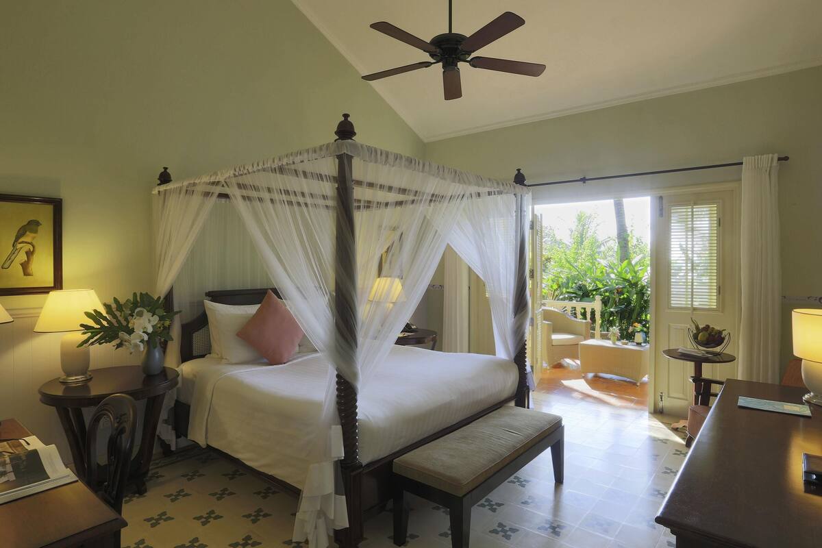 A four-poster bed with a canopy, a ceiling fan, a bedside table with a lamp, a chair, and a view of the outdoors.