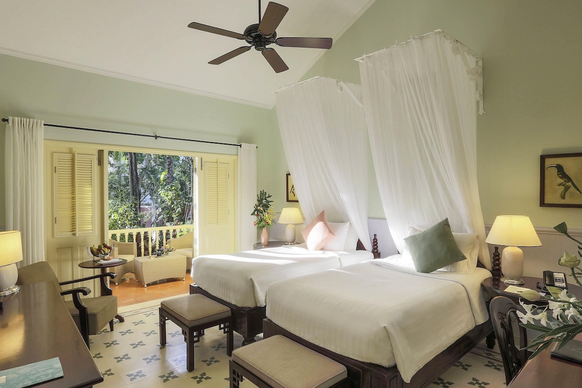 A hotel room with two beds, a ceiling fan, a balcony with a view of greenery, and a desk with a chair.