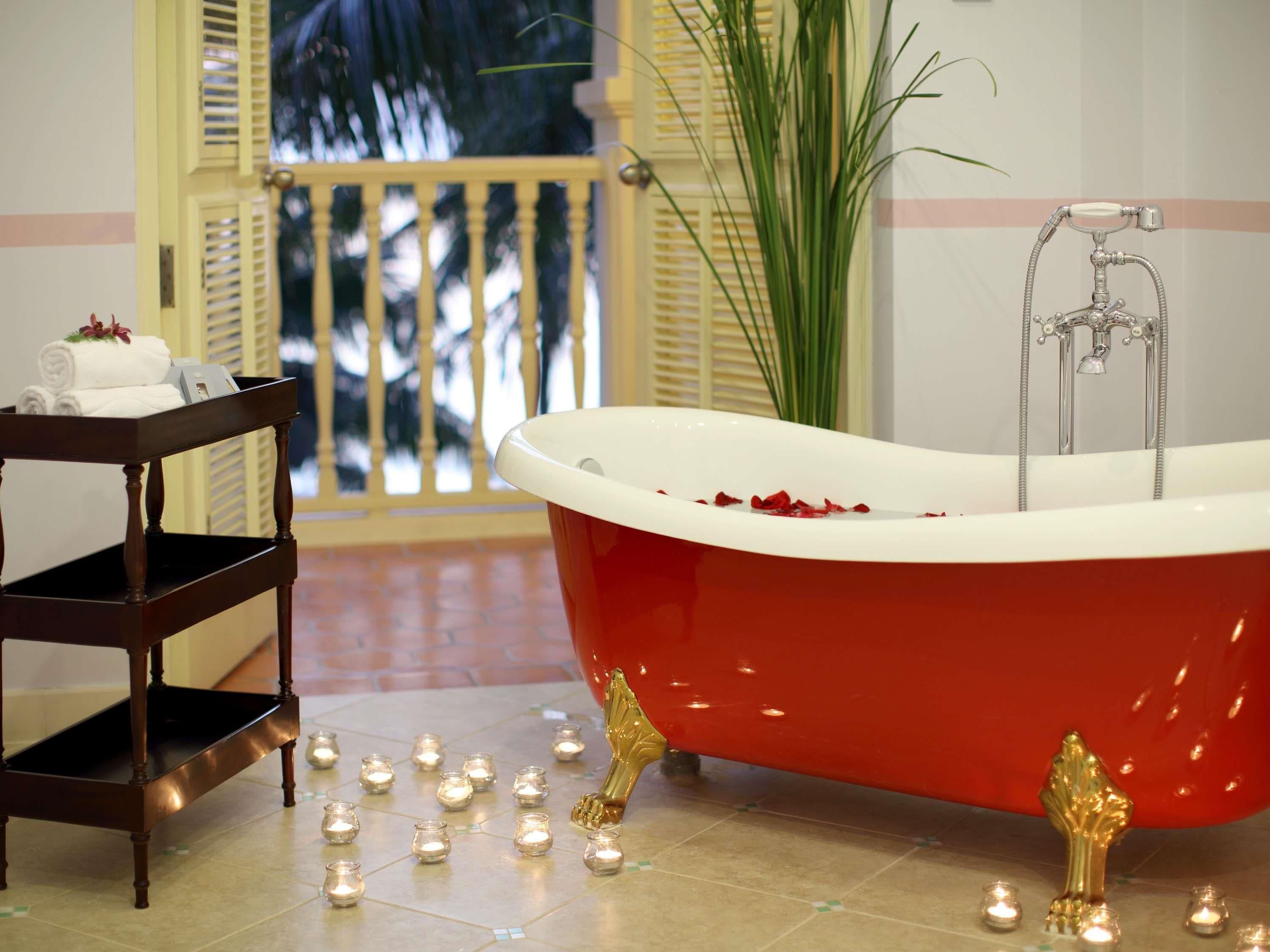 Une baignoire rouge avec un pommeau de douche, une table d’appoint en bois avec des serviettes et des bougies posées sur le sol.