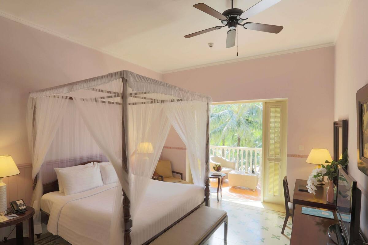 A four-poster bed with a canopy, a ceiling fan, a desk with a lamp, and a television.