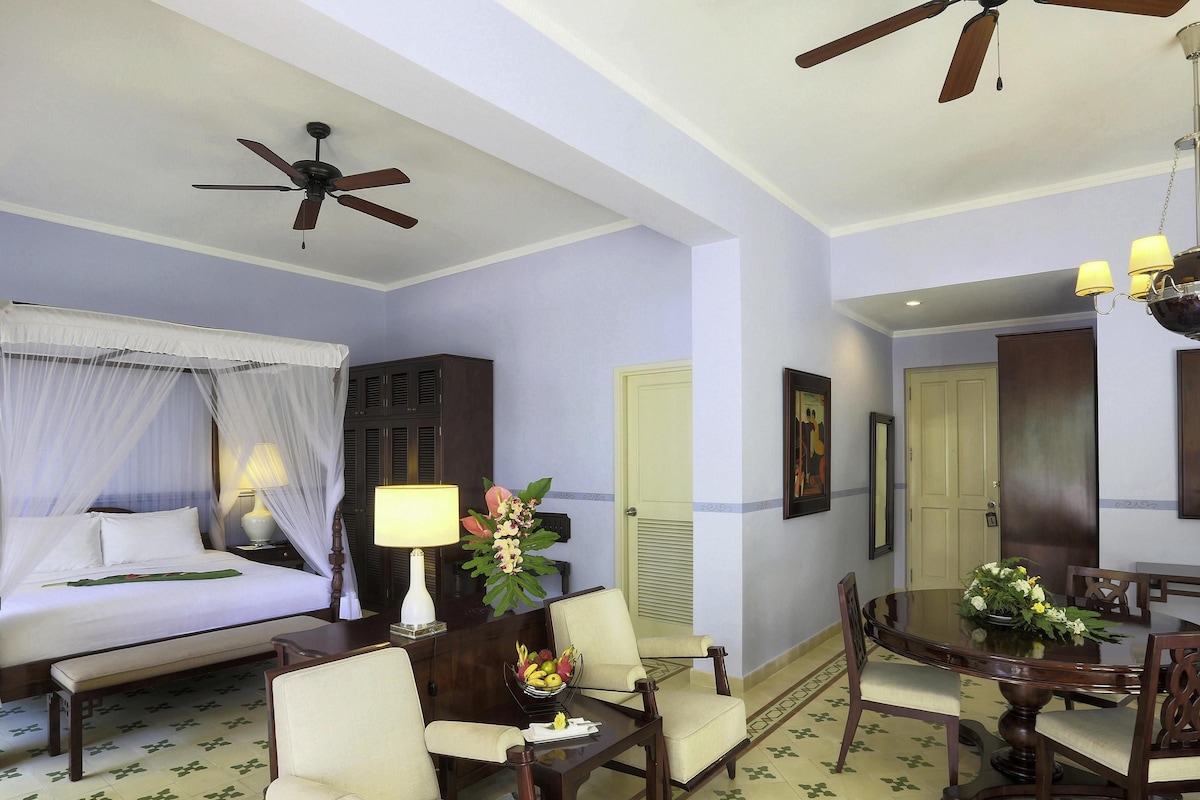 A hotel room with a bed, a desk, a dining table, chairs, a ceiling fan, and a door.