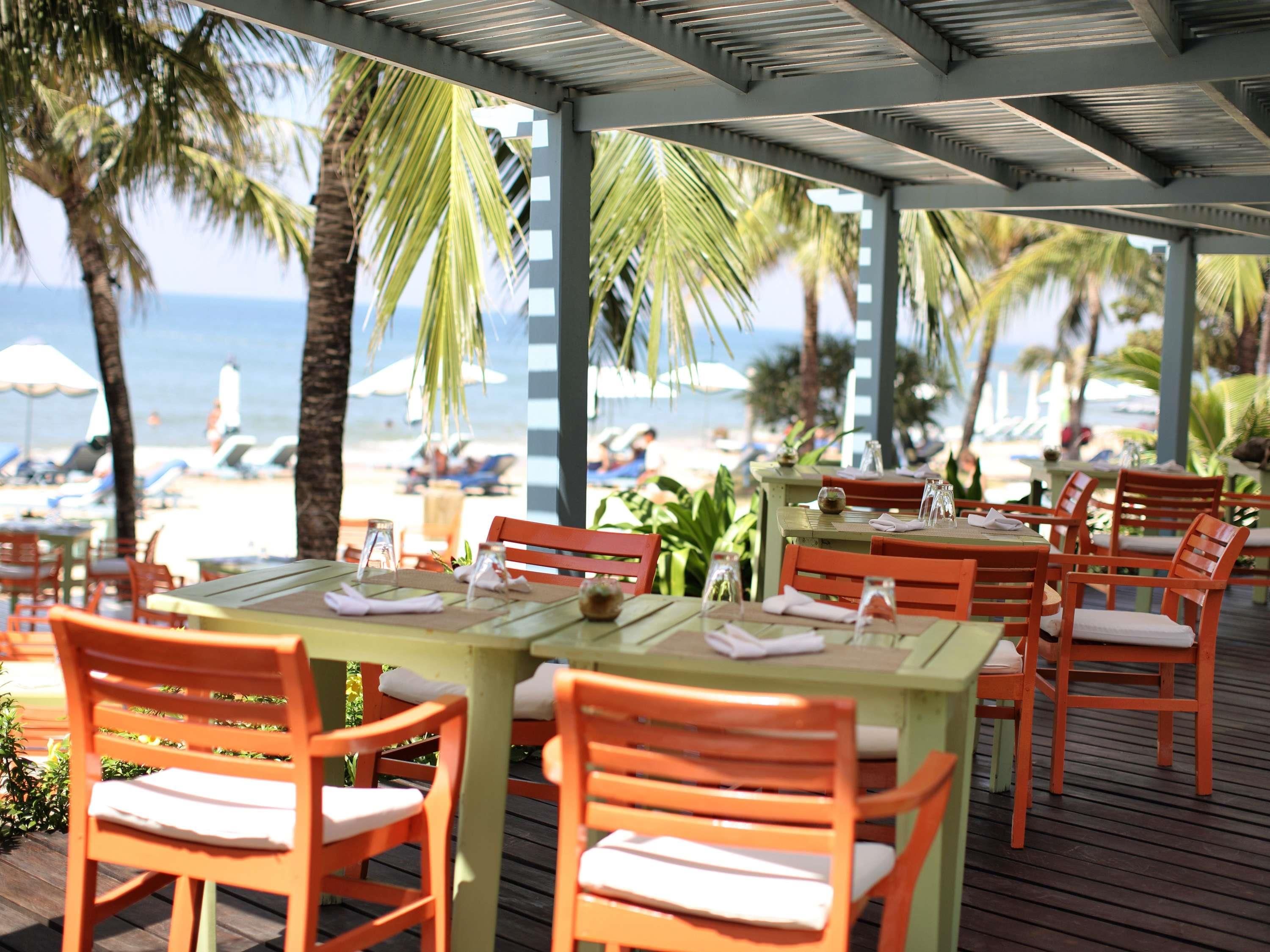Un área de comedor al aire libre con mesas y sillas de madera, con vista a una playa con palmeras y sombrillas.