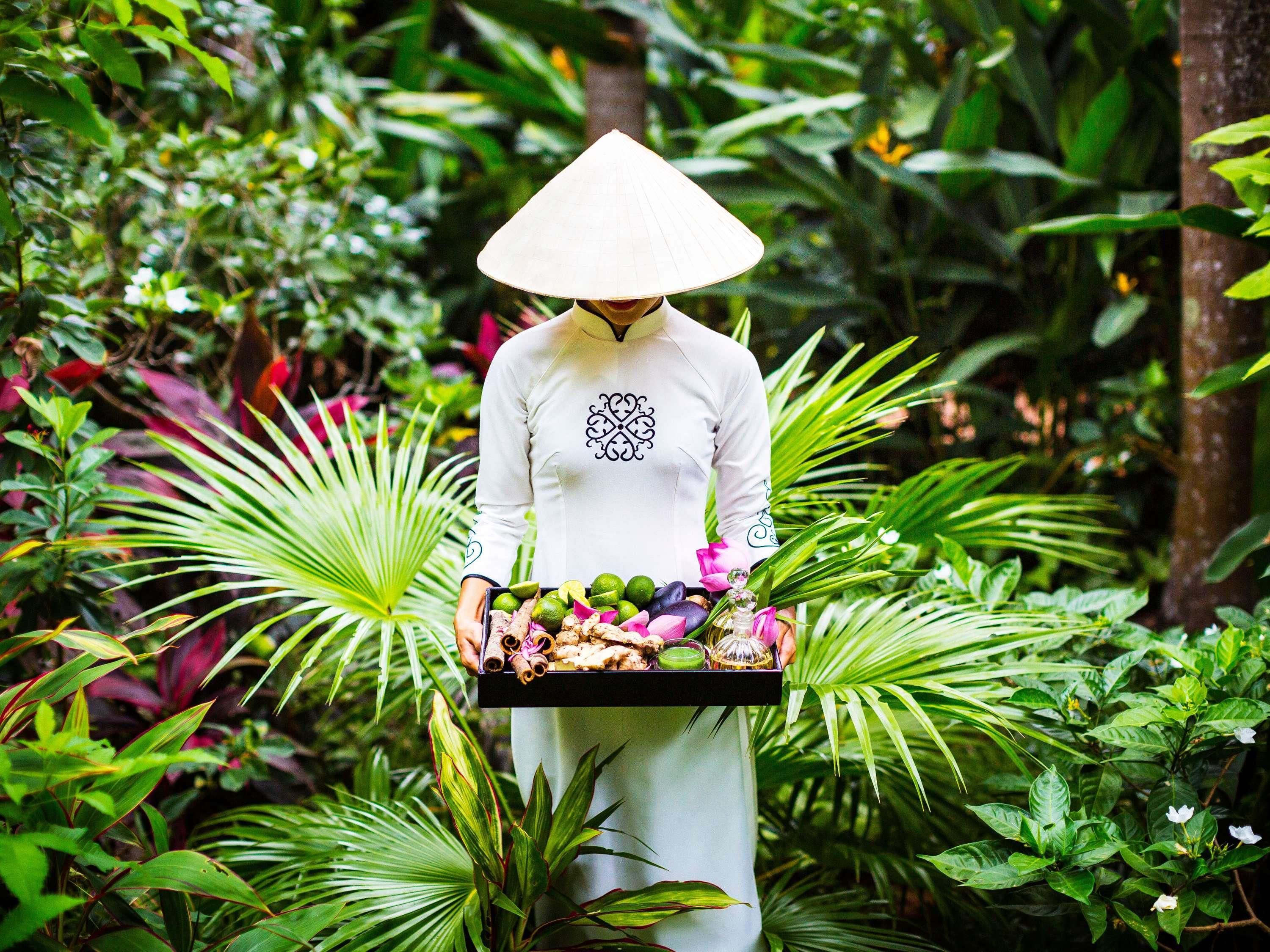 A person in a white traditional outfit carrying a tray with various fruits and flowers in a lush, green environment.