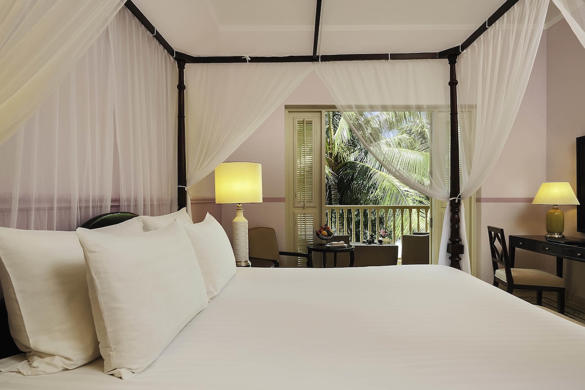 A four-poster canopy bed with white linens, a small table with a lamp, and a view of palm trees through a window.