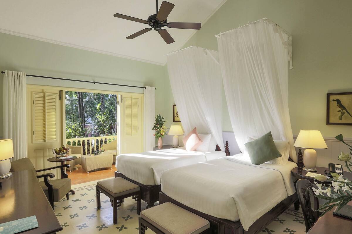Ein Hotelzimmer mit zwei Betten, einem Deckenventilator, einem Balkon mit Blick auf Grünflächen und einem Schreibtisch mit Stuhl.