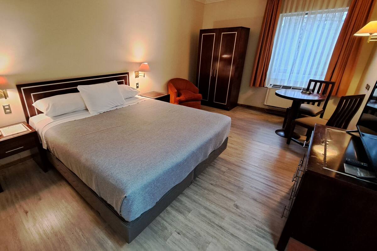 Une chambre d’hôtel avec un grand lit, des tables de chevet, un bureau, une chaise, une armoire et une fenêtre avec des rideaux.