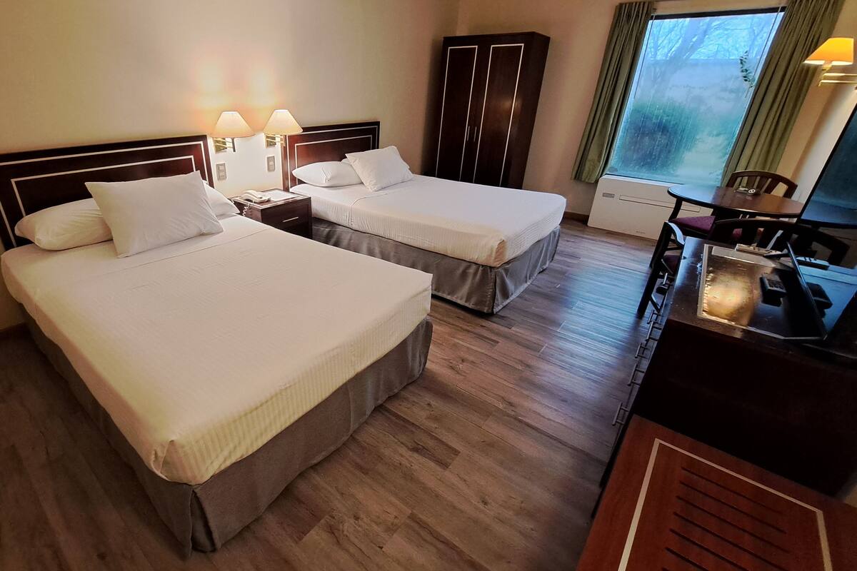 Une chambre d’hôtel avec deux lits, un plancher en bois, un bureau avec une lampe et une fenêtre avec des rideaux.