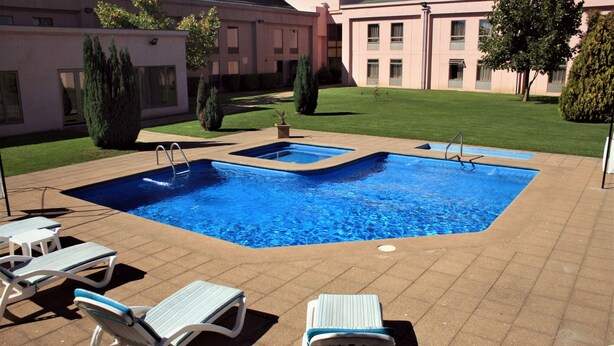 A swimming pool with two sections, surrounded by lounge chairs and a building in the background.