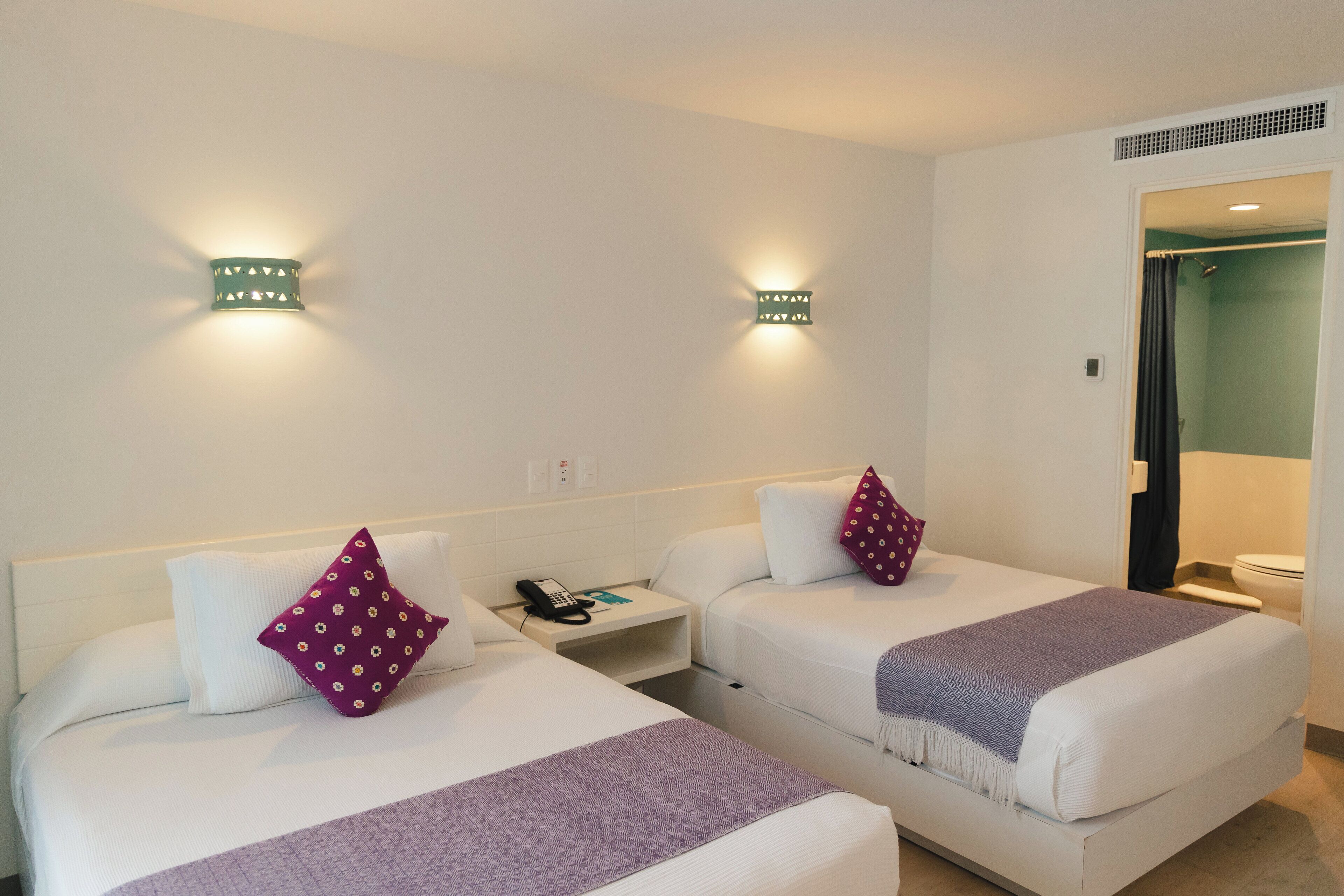 Habitación de hotel con dos camas, cada una con almohada y manta, mesita de noche con teléfono y lámparas de pared.