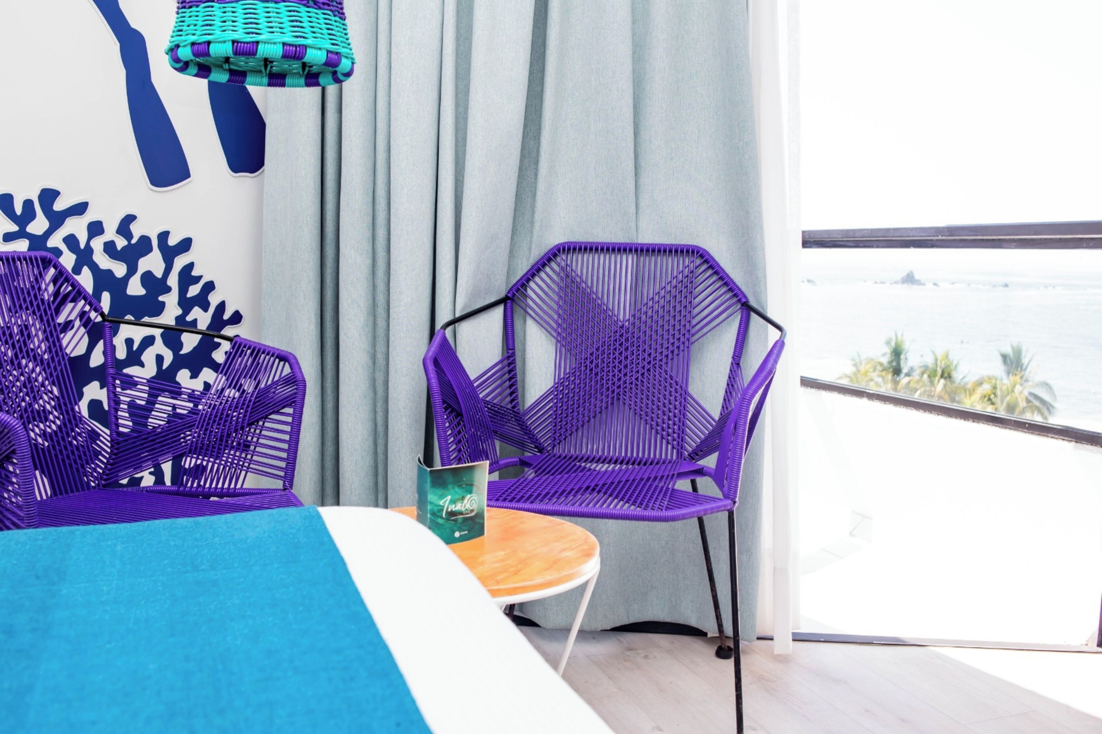A room with a bed, two purple chairs, a small table, and a view of the ocean.