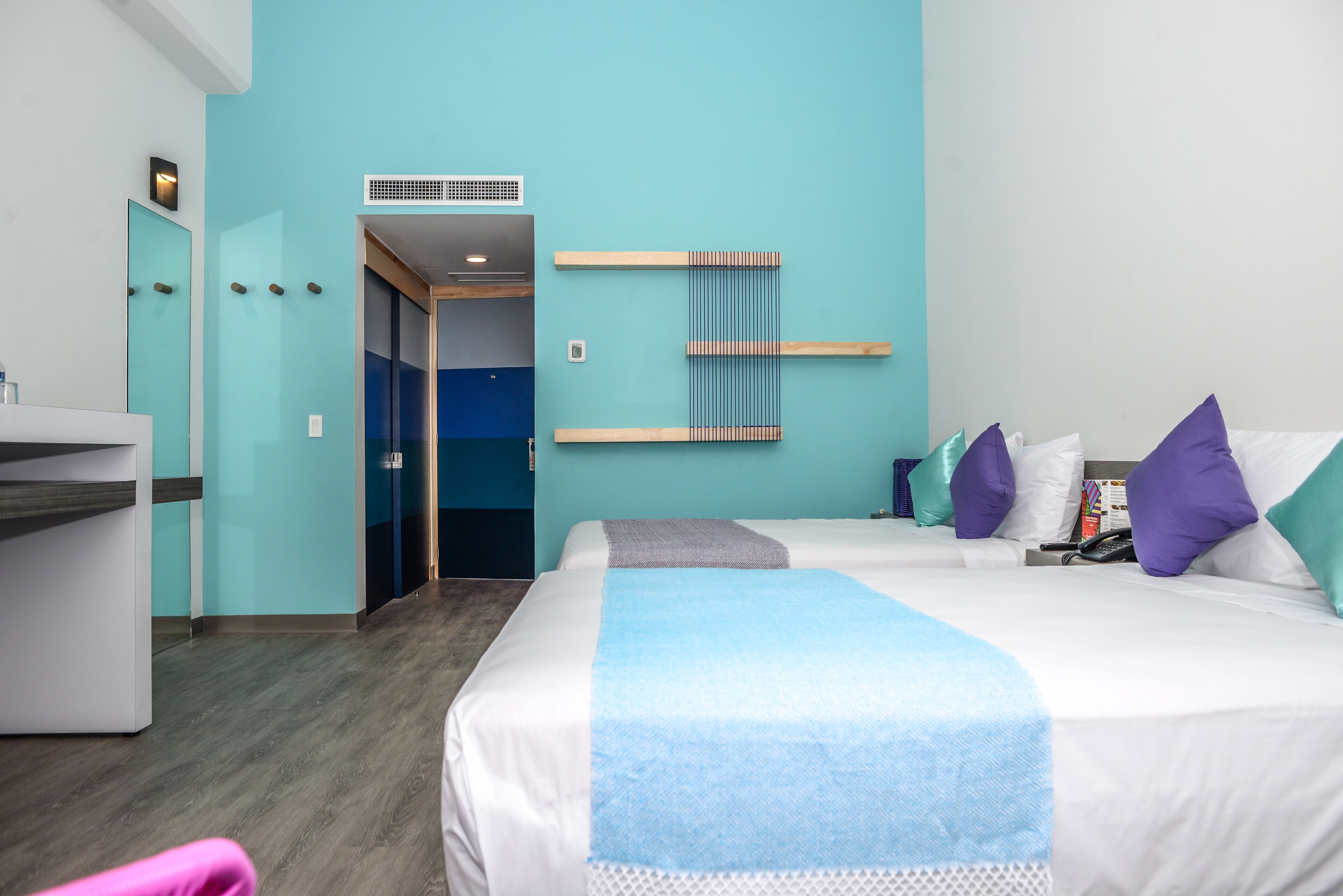 A hotel room with a teal wall, two beds with white and blue bedding, a small desk, and a bathroom visible through a glass door.