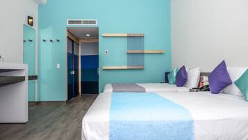 A hotel room with a teal wall, two beds with white and blue bedding, a small desk, and a bathroom visible through a glass door.