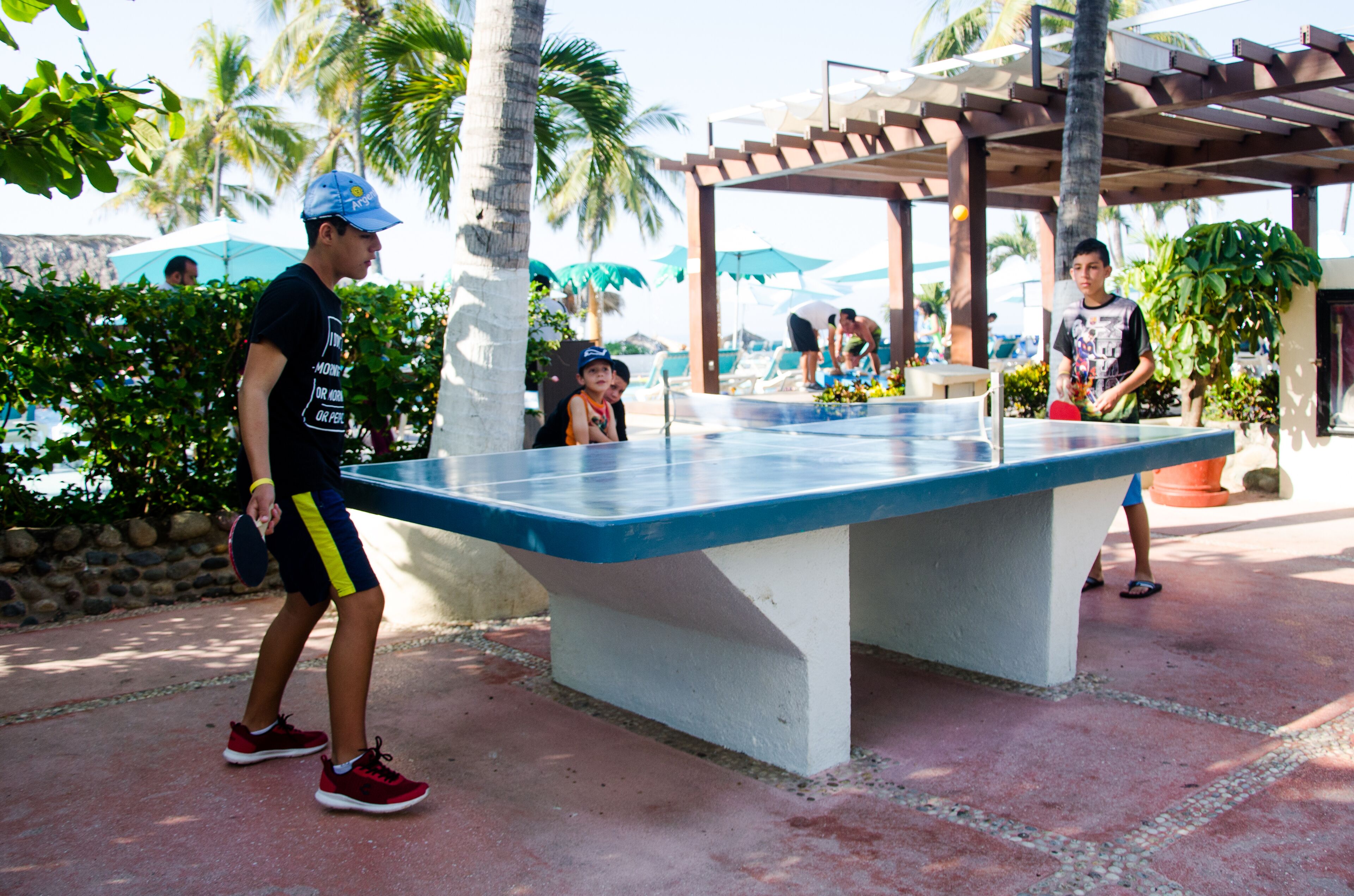Una mesa de ping pong al aire libre en una zona de resort, con palmeras y sombrillas al fondo.