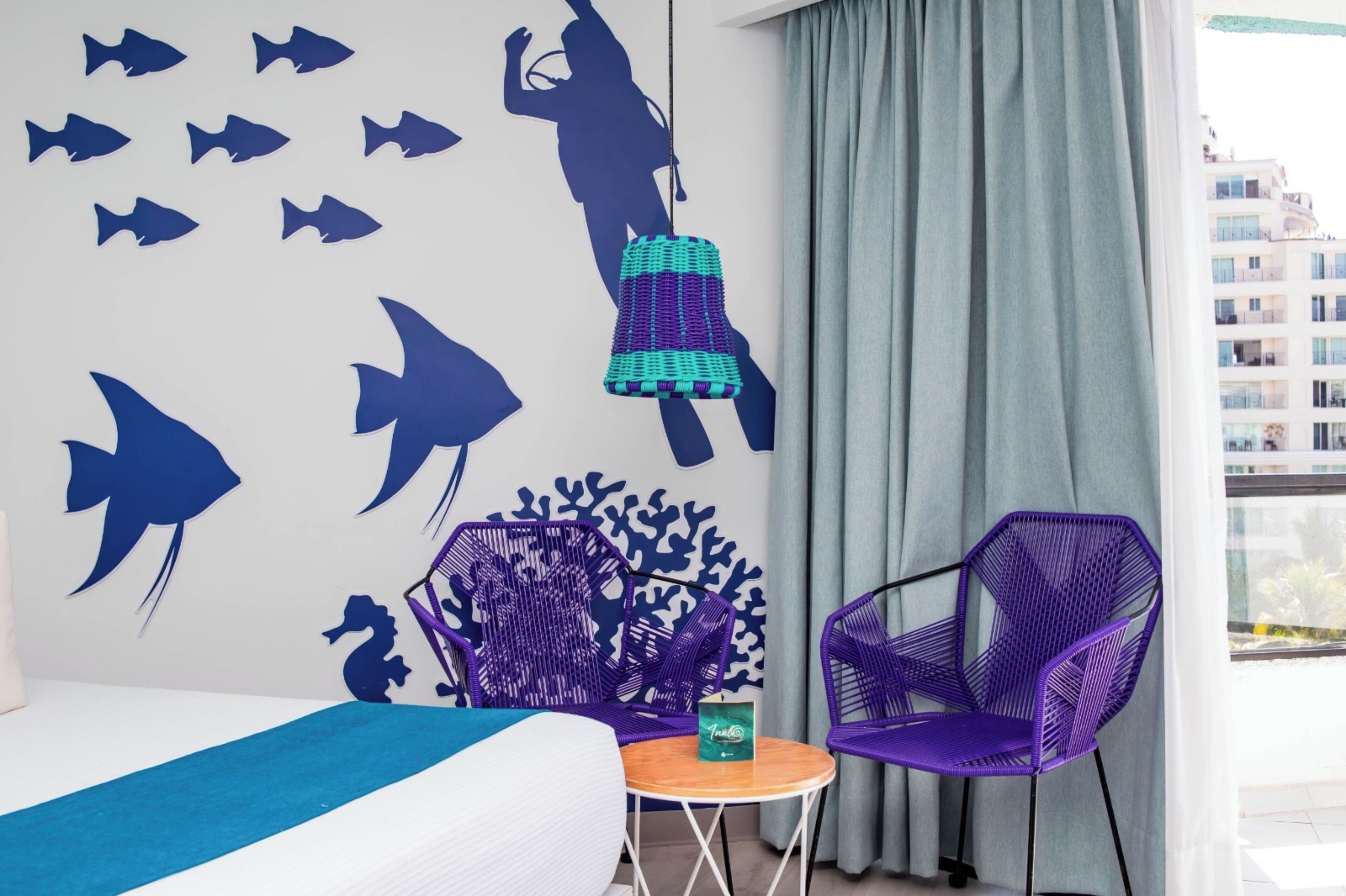 A hotel room with a bed, two purple chairs, a small table, a wall with fish and seahorse decals, and a view of buildings through a window.
