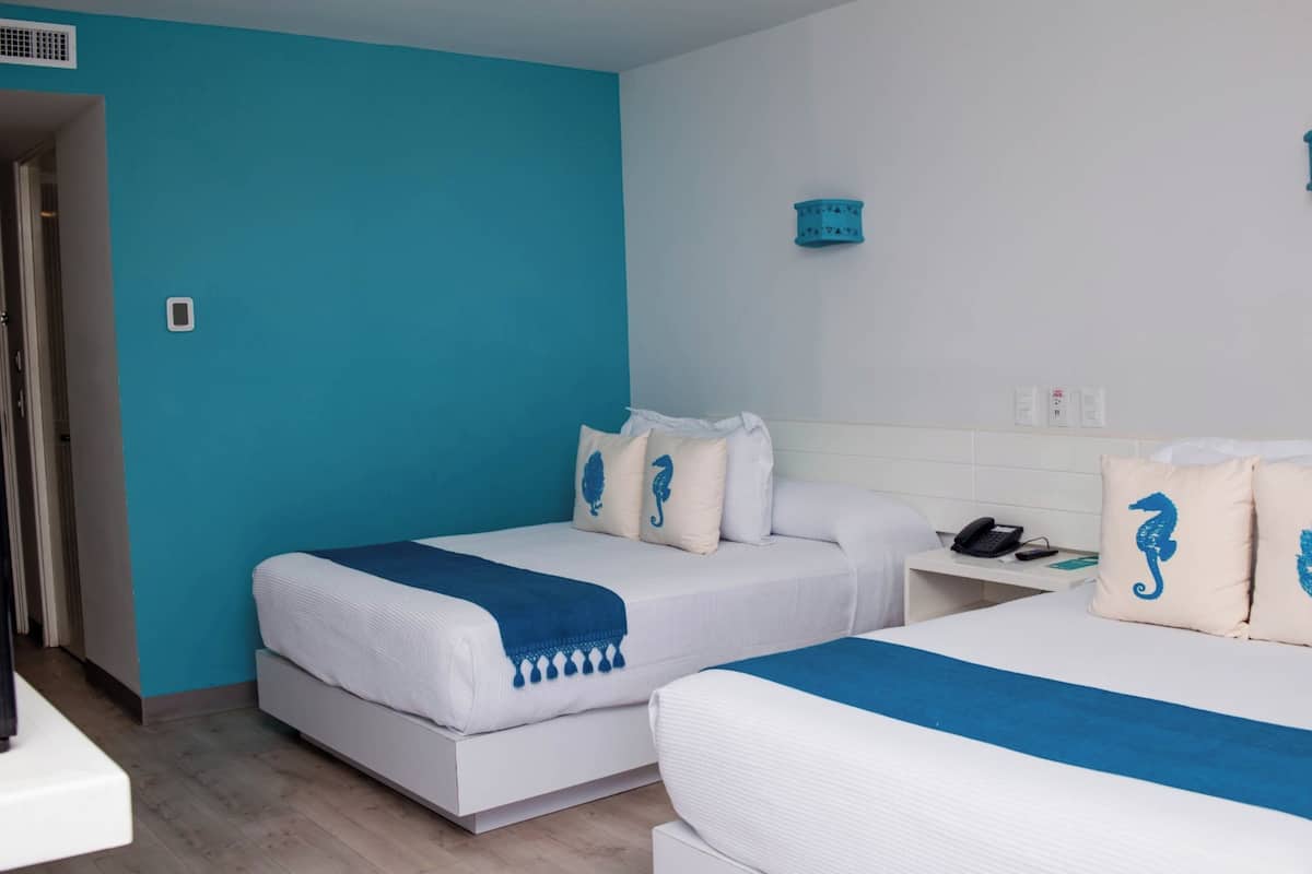 Hotellihuone, jossa on kaksi sänkyä, valkoinen yöpöytä, jossa on puhelin, ja sininen tehosteseinä.