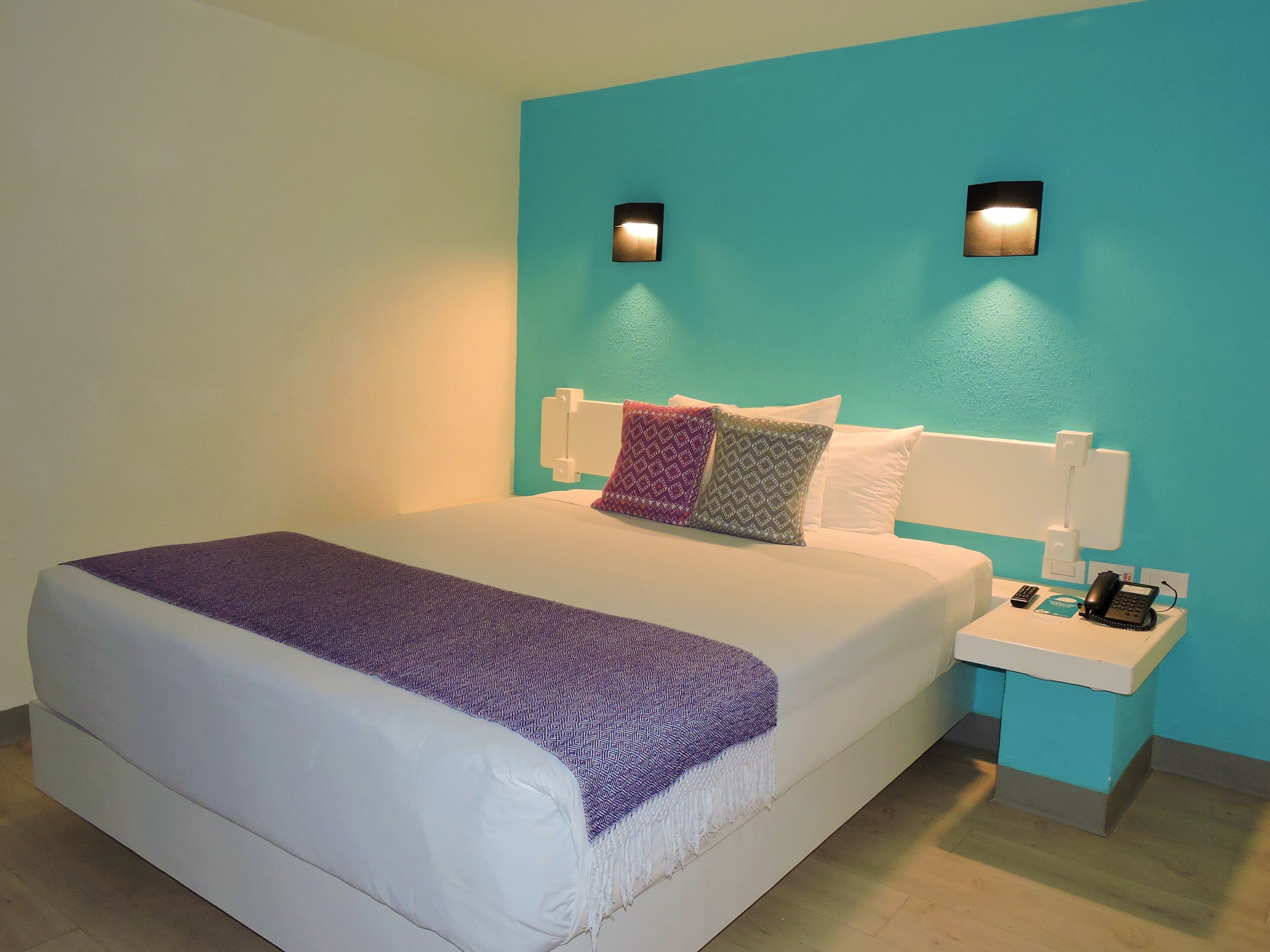 A neatly made bed with a white mattress and a purple blanket, a nightstand with a phone and a lamp, and teal walls with two wall-mounted lights.
