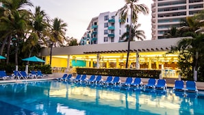 A swimming pool with blue lounge chairs, a restaurant with yellow lighting, and palm trees. - Fontan Ixtapa Beach Resort - All Inclusive (Ixtapa)