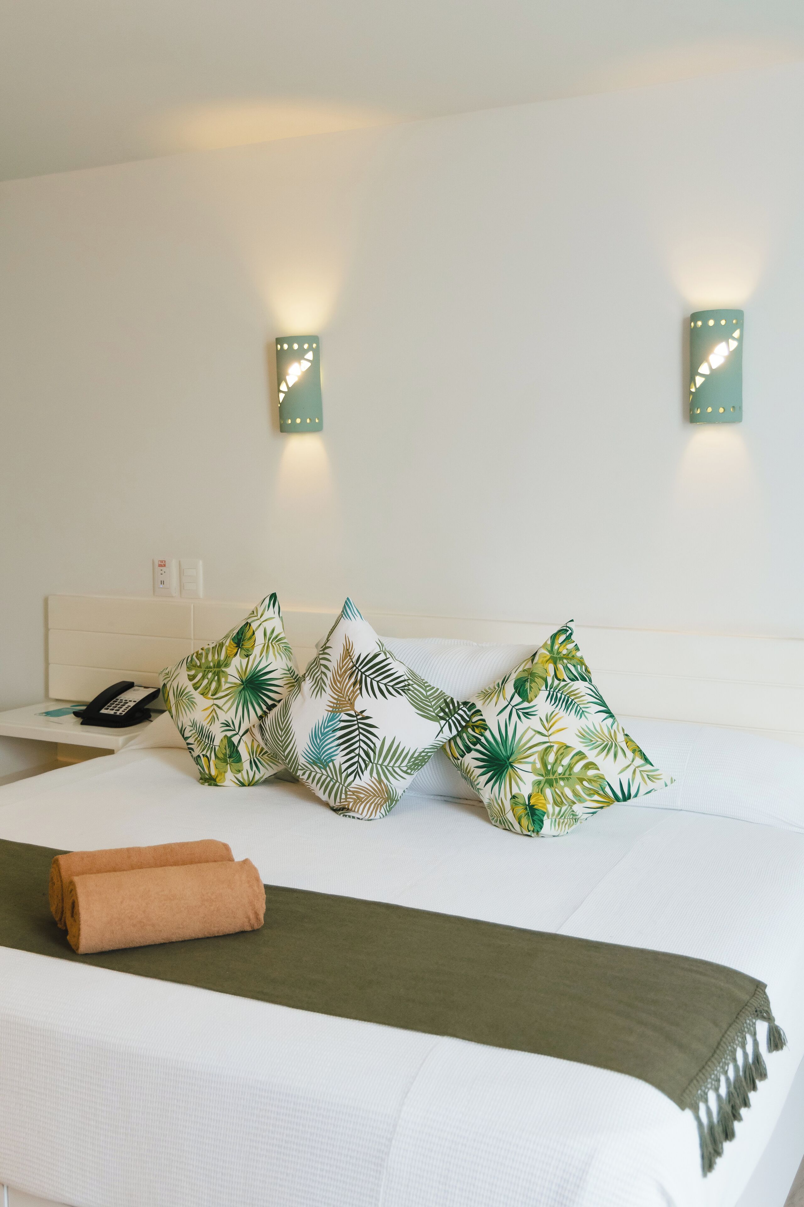A neatly made bed with a green throw blanket, two rolled towels, and three pillows with a tropical leaf pattern. There are two wall-mounted lights and a telephone on a bedside table.
