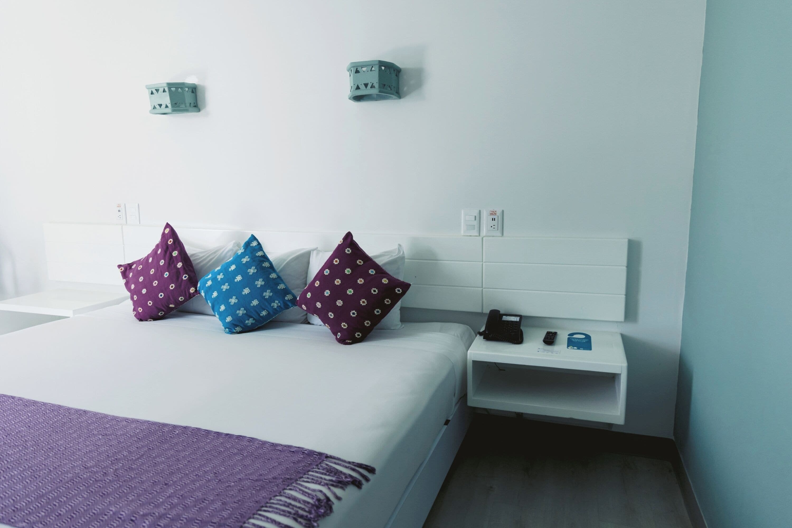 A hotel room with a bed, pillows, a nightstand, a phone, and a wall socket.