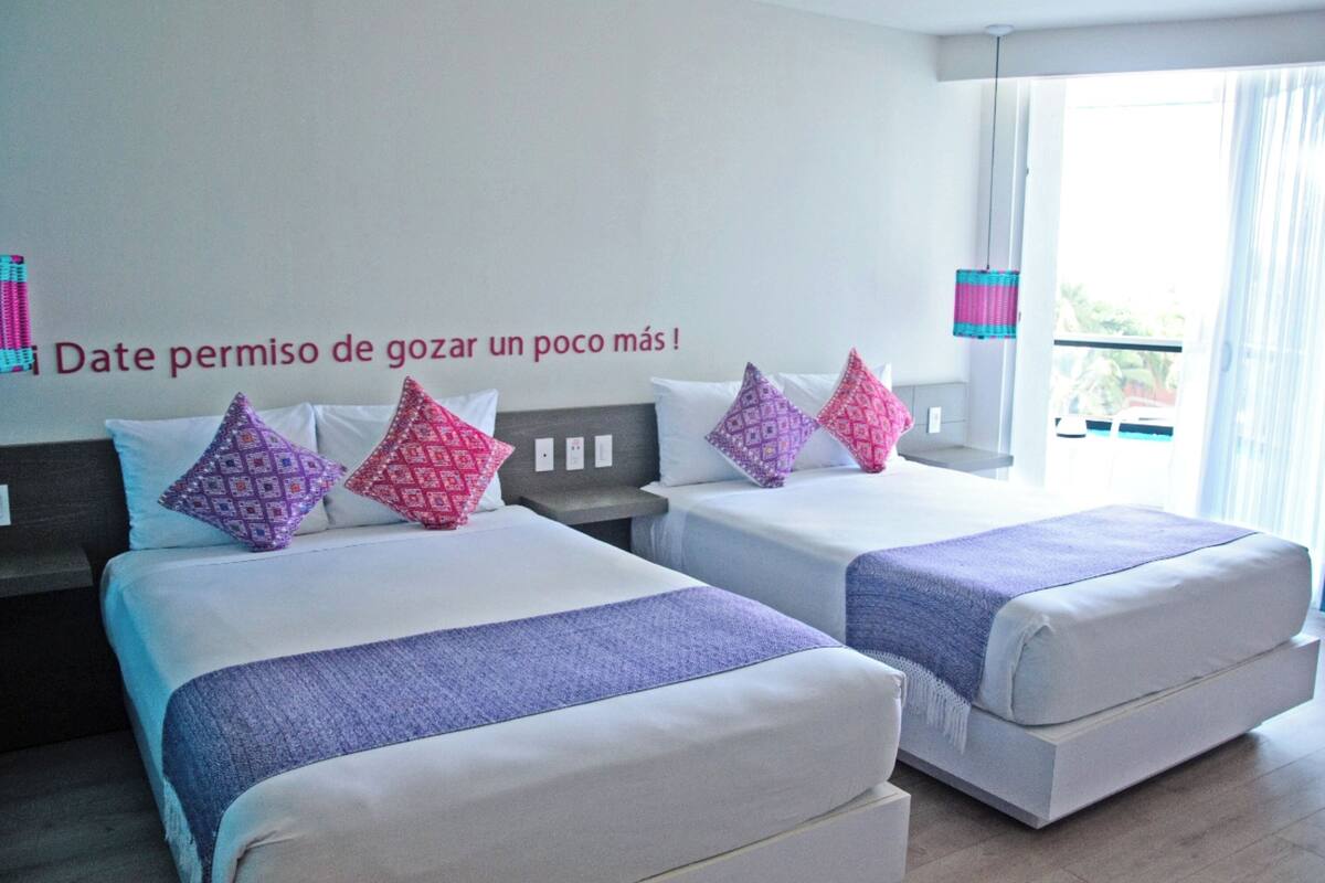 Hotellihuone, jossa on kaksi sänkyä, ja jokaisessa sängyssä on kuviollisia tyynyjä ja violetti peitto.