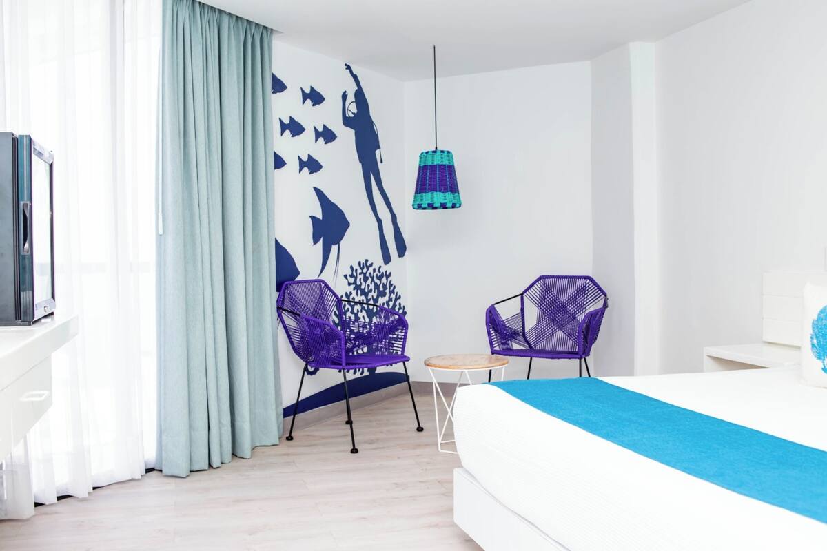 Moderni hotellihuone, jossa on sänky, kaksi violettia tuolia, pieni pöytä, seinämaalaus, jossa on kuva sukeltajasta, ja riippuvalaisin.
