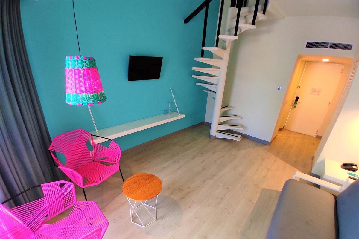 A room with teal walls, a wooden floor, a staircase, a small table with a stool, and two pink chairs.