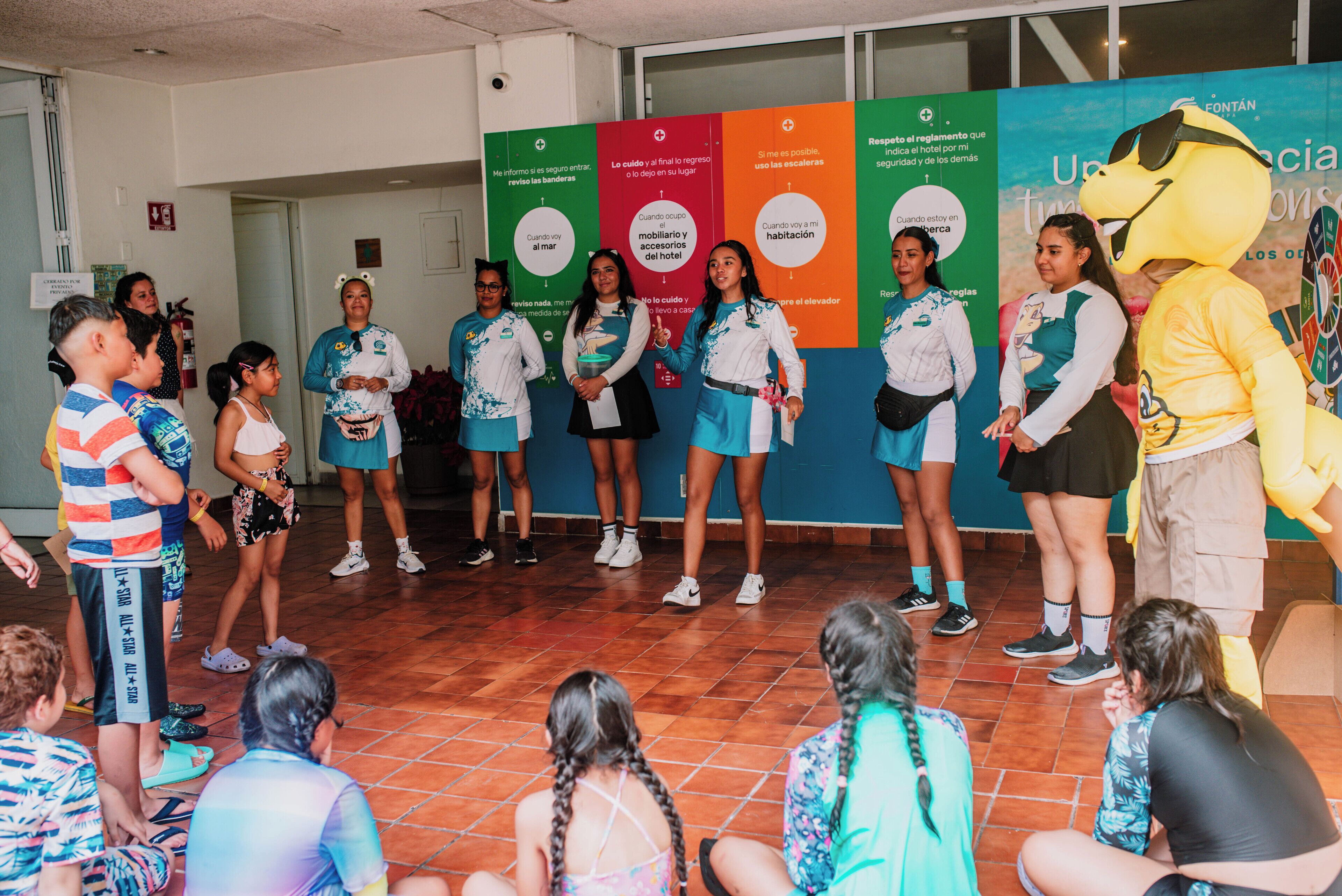 A group of people in a room with a tiled floor, standing and sitting in front of a wall with colorful panels.