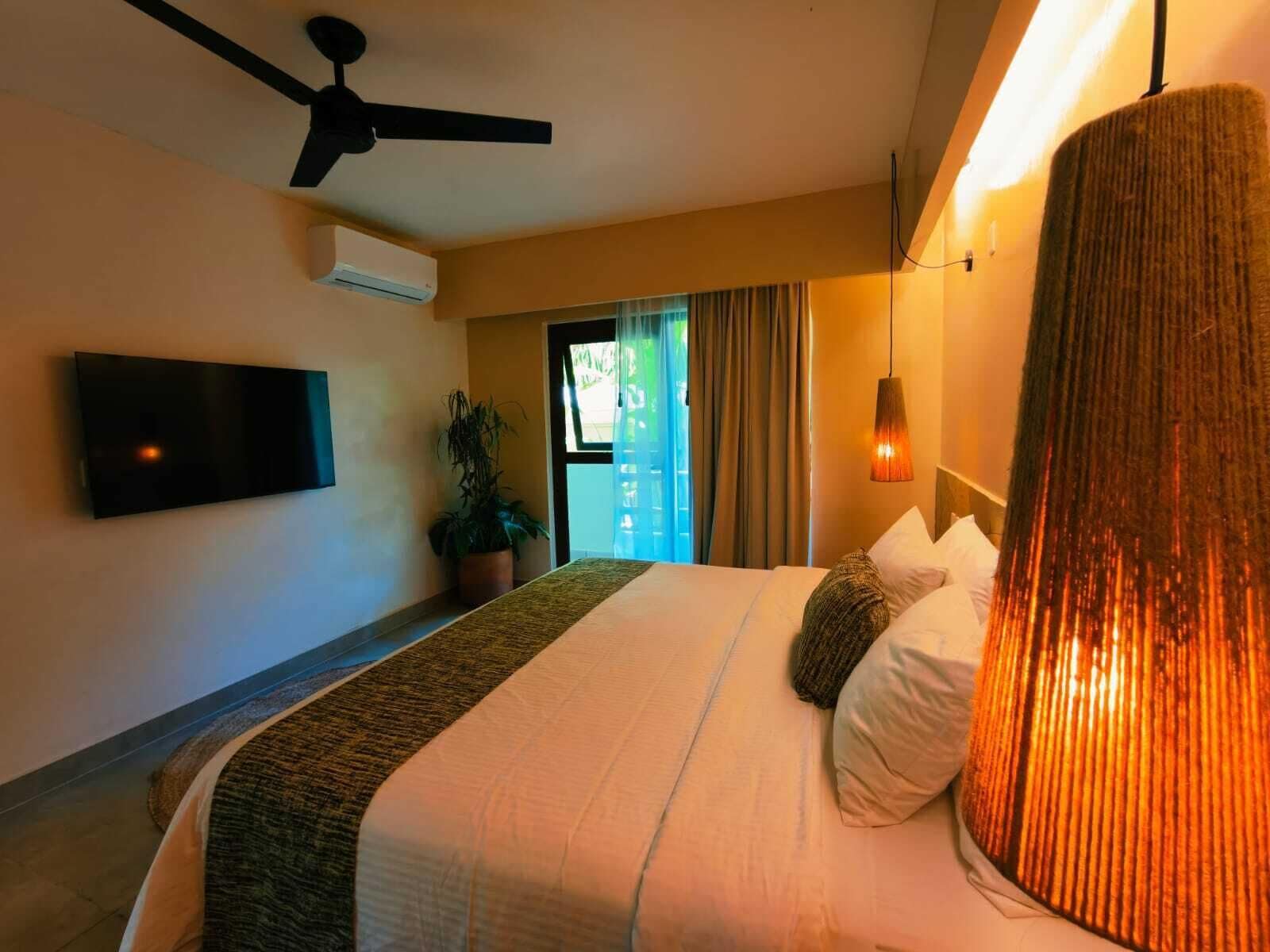 Habitación de hotel con cama, ventilador de techo, equipo de aire acondicionado, televisor, planta en maceta y vista al exterior.