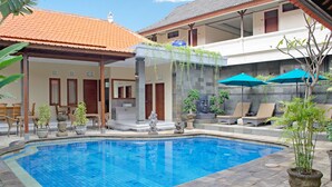A swimming pool with tiled flooring, surrounded by lounge chairs and umbrellas, and a covered seating area with chairs and potted plants. - Hotel Flamboyan (Kuta)