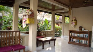 A covered outdoor seating area with a wooden bench and table, surrounded by plants and a bookshelf. - Hotel Flamboyan (Kuta)