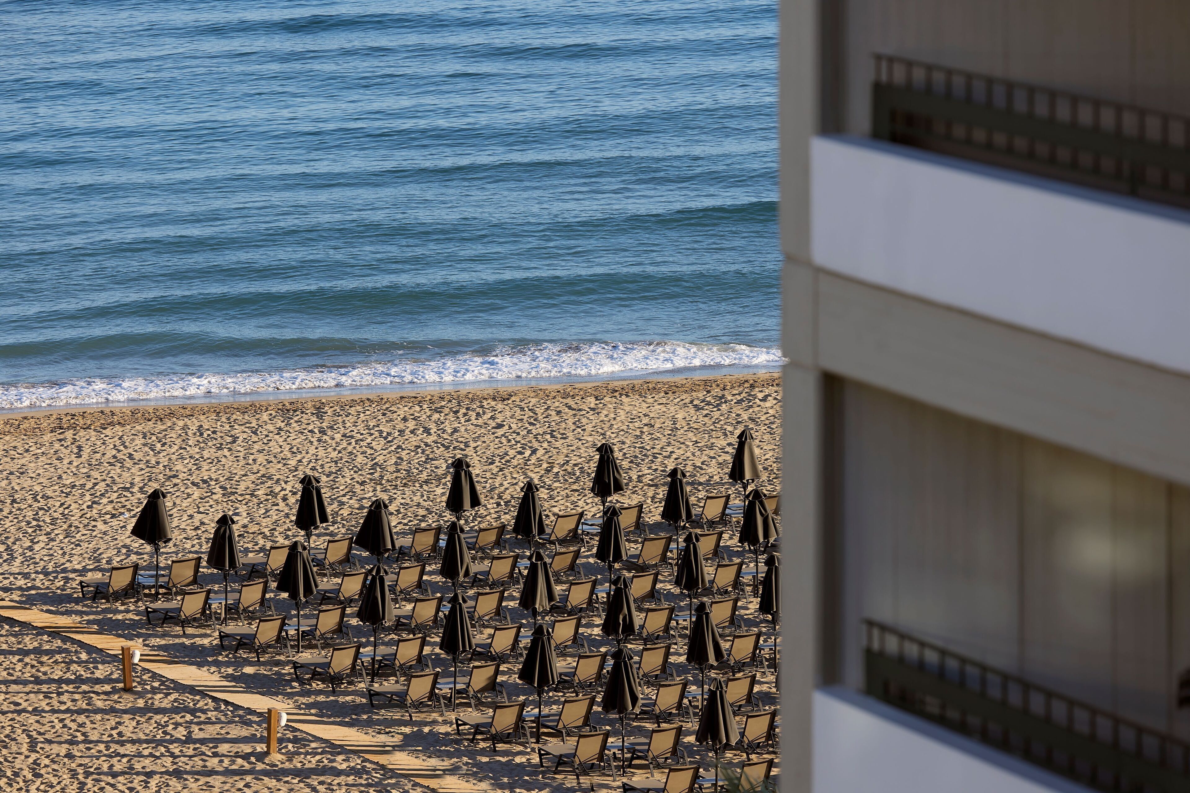 A beach with rows of sun loungers and umbrellas, a building in the foreground, and the ocean in the background.