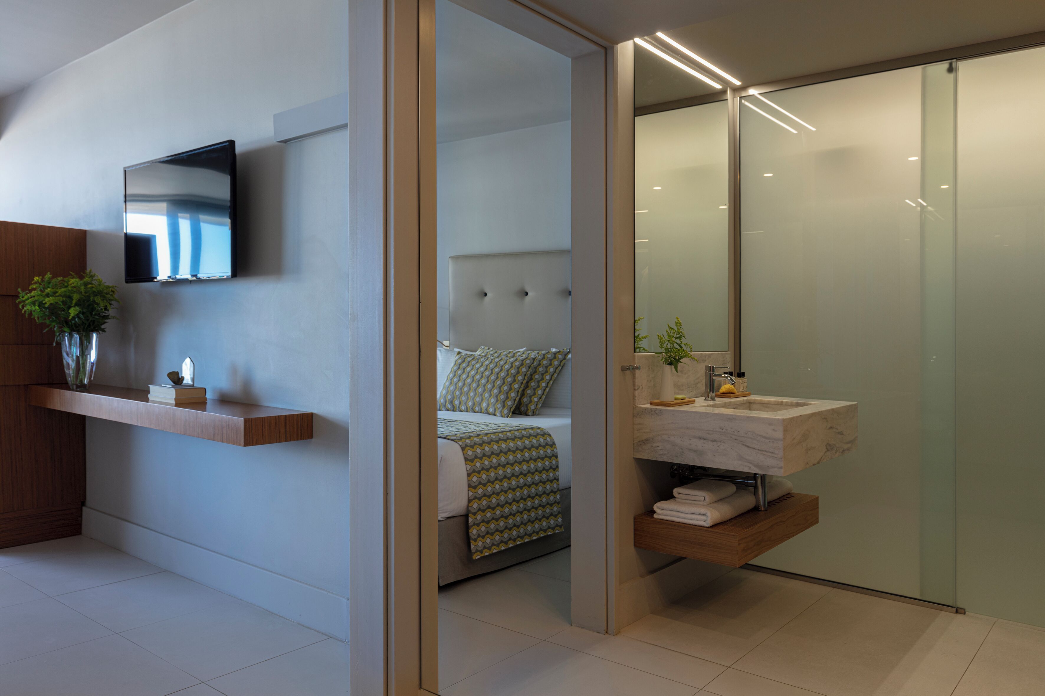 A modern hotel room with a bed, a TV mounted on the wall, a wooden shelf with a plant, and a glass shower enclosure.