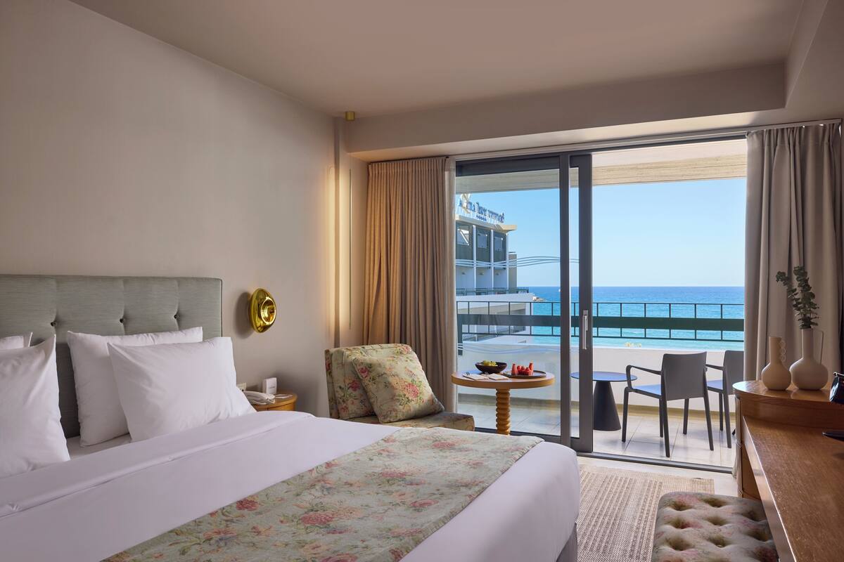 Quarto de hotel com cama, uma cadeira, uma mesinha com frutas e uma varanda com vista para o mar.