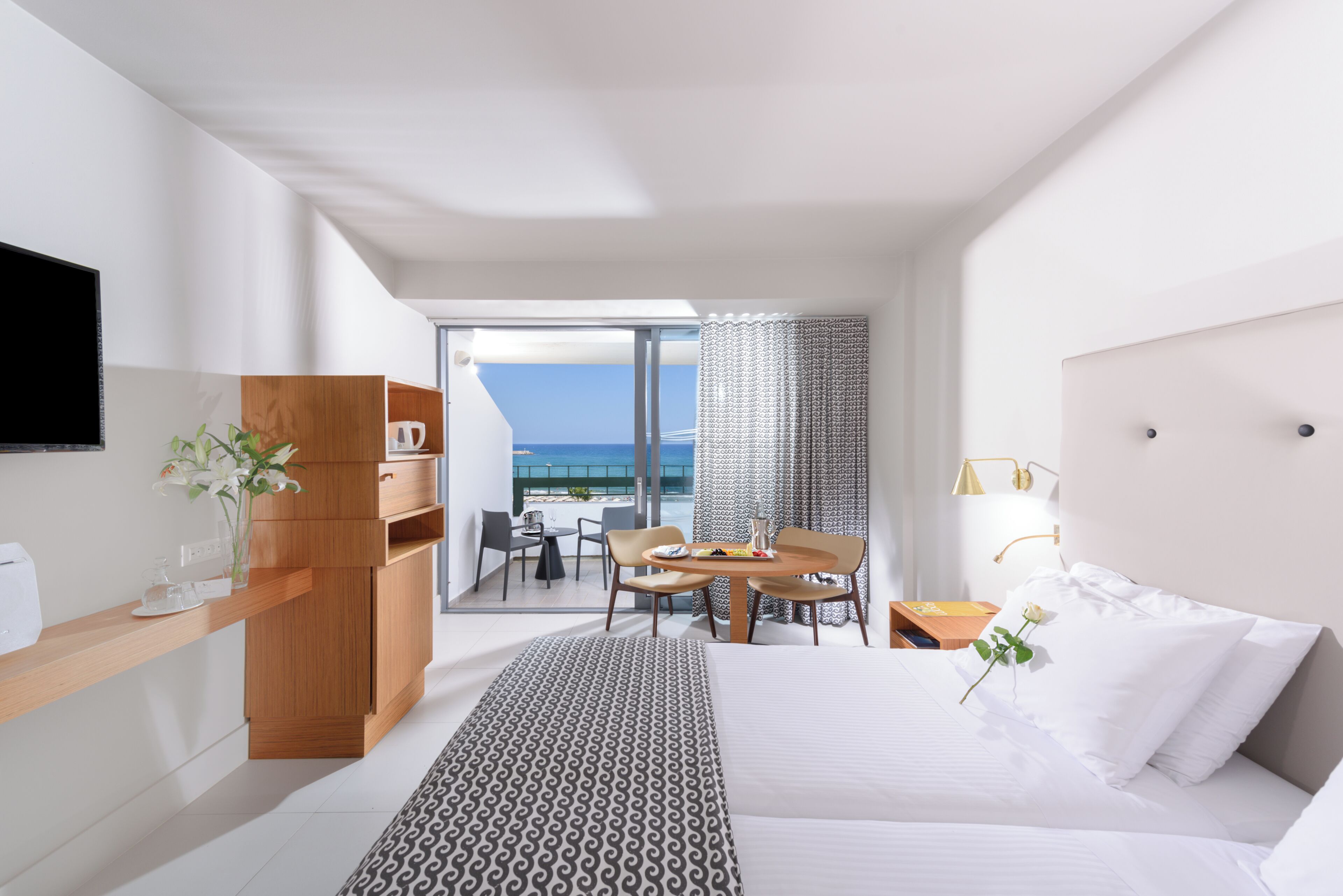 Une chambre d’hôtel moderne équipée d’un lit, d’un bureau avec un ordinateur, d’un coin repas avec une table et des chaises, et d’un balcon offrant une vue sur la mer.