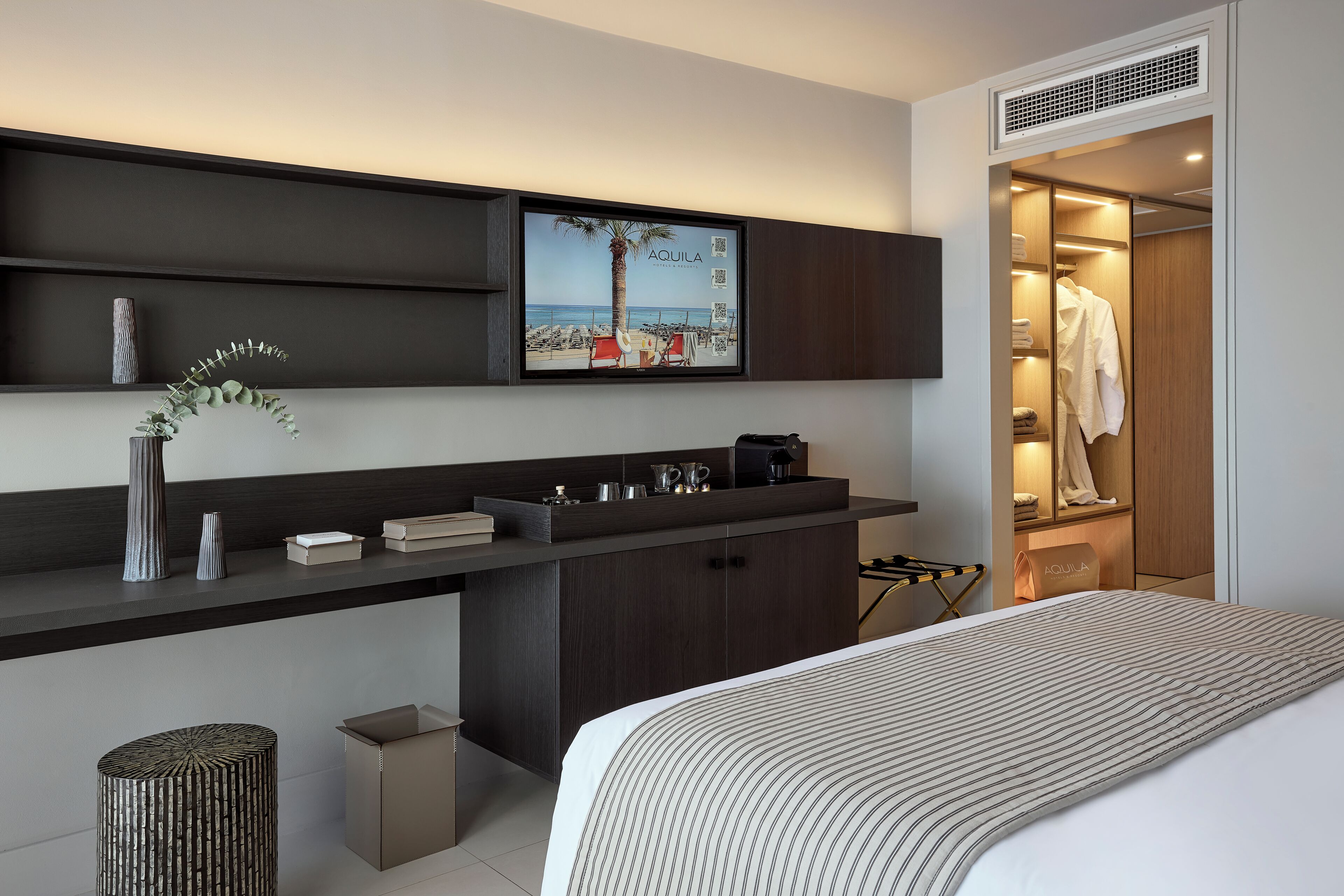 A modern hotel room with a bed, a desk with a flat-screen TV, a wardrobe, and a small trash bin.