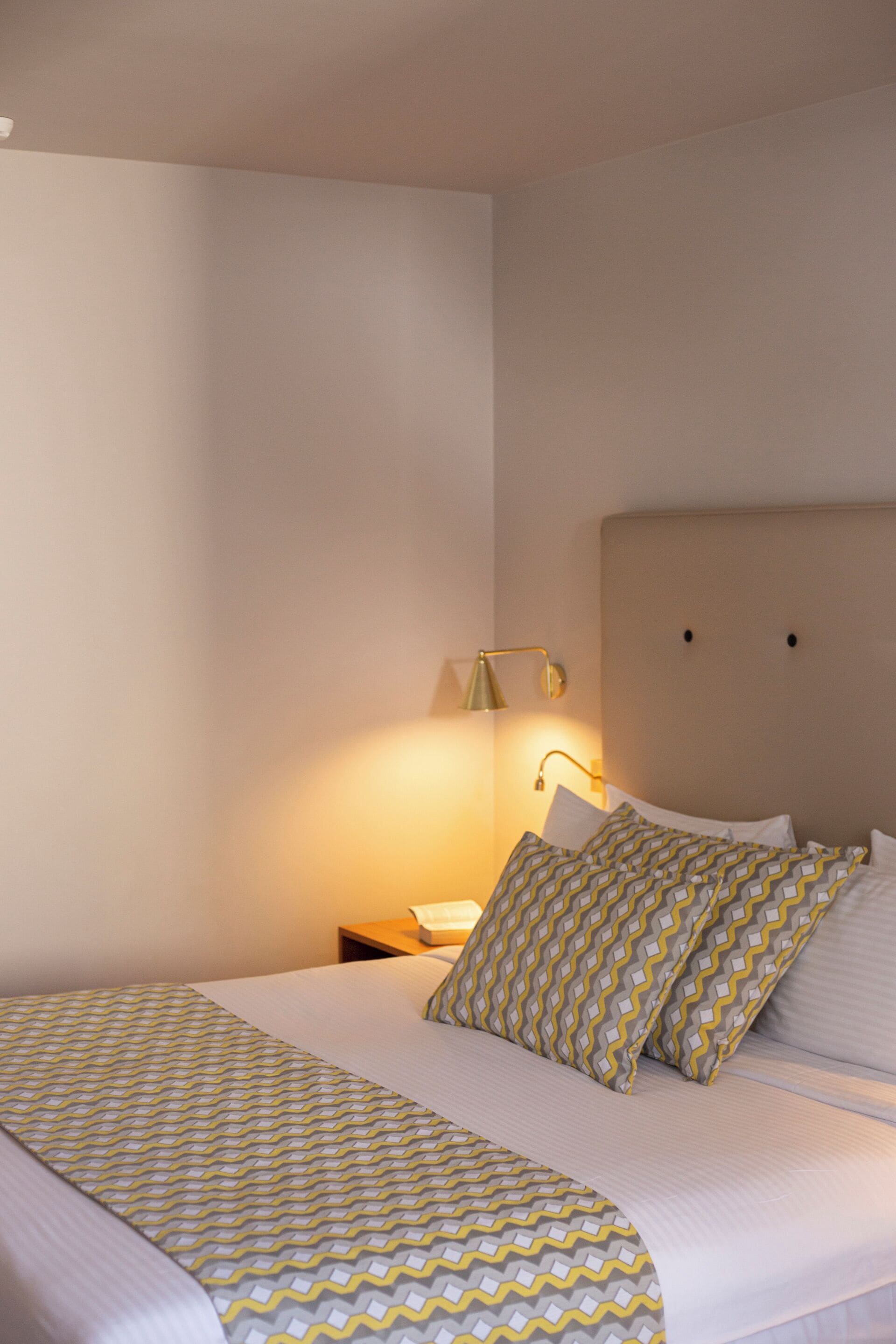 A neatly made bed with patterned bedding, a bedside lamp, and a plain wall.