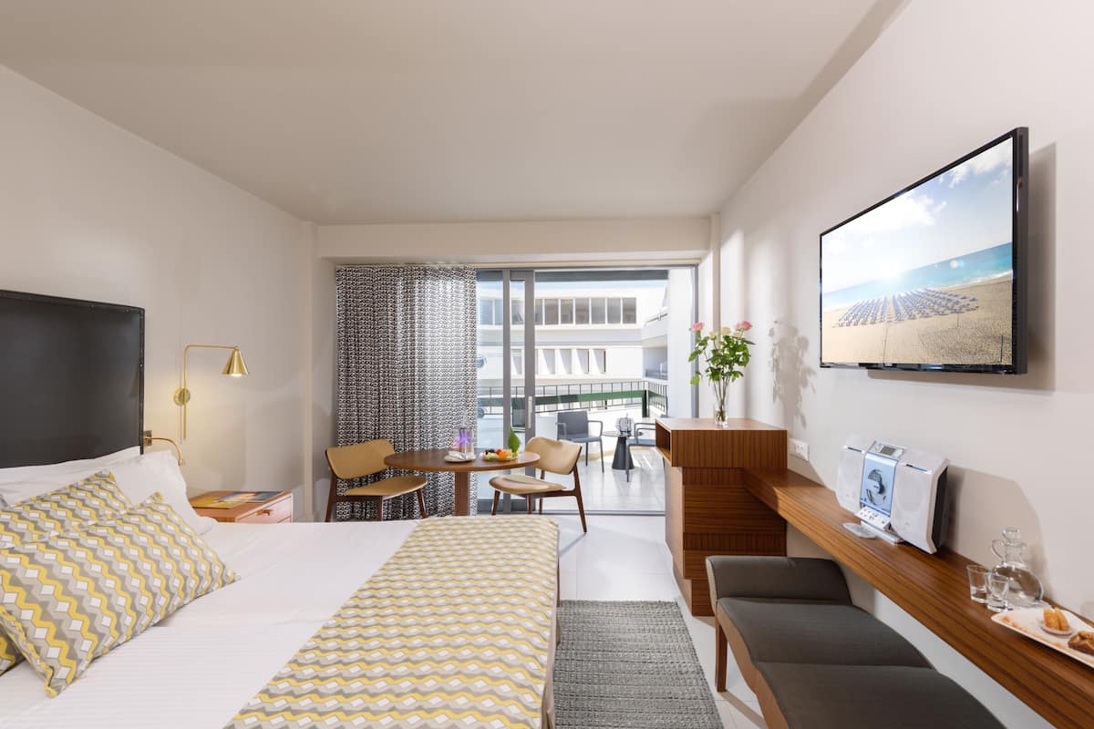 Quarto de hotel moderno com cama, escrivaninha, cadeira, máquina de café e televisão na parede.