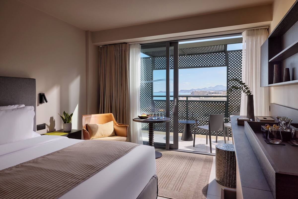 Quarto de hotel com cama, área de estar, mesinha e varanda com vista.