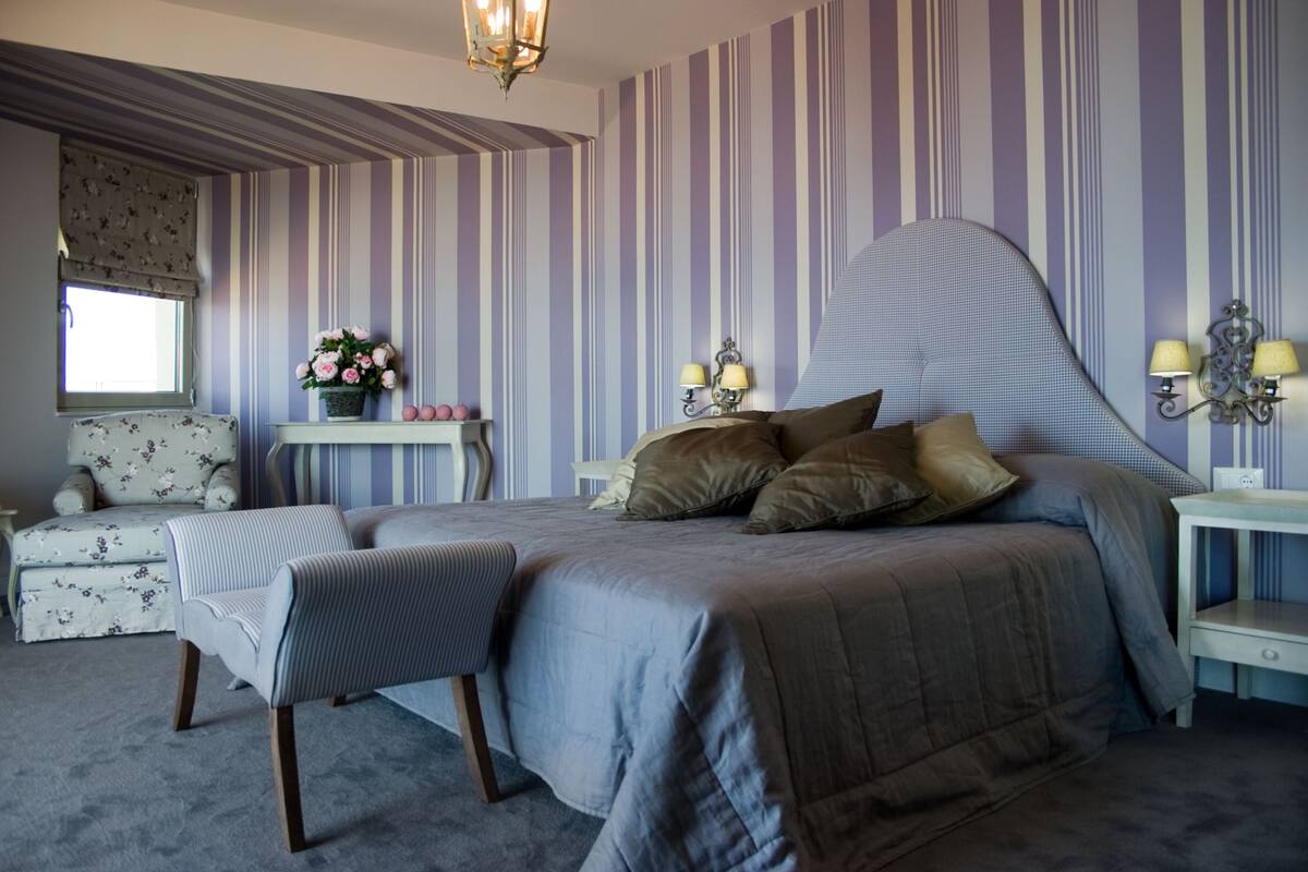 Quarto com uma cama grande, mesa de cabeceira, cadeira, pequena mesa com flores e janela com cortinas.