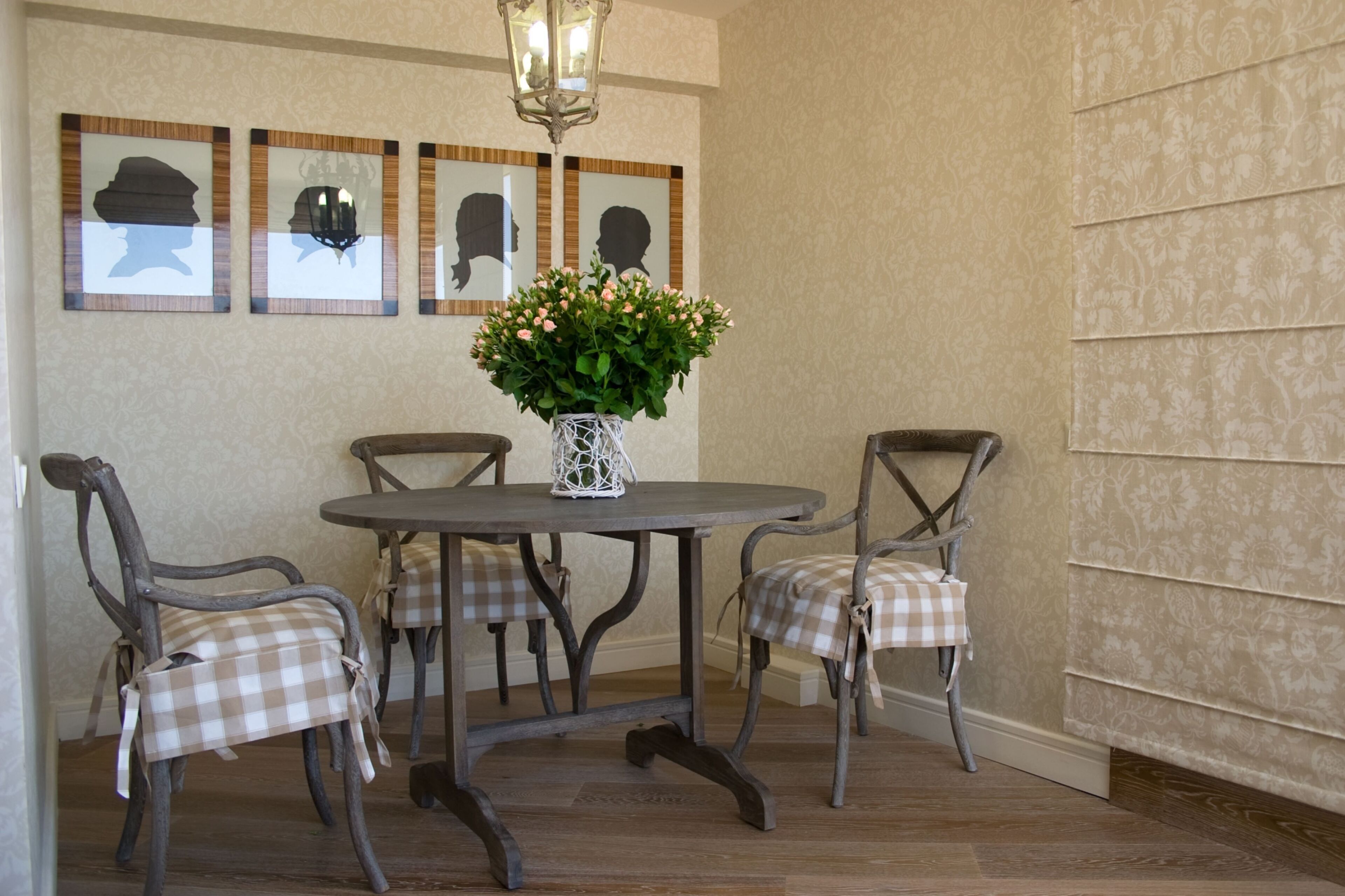 A dining area with a round table, four checkered chairs, a vase with flowers, and framed silhouettes on the wall.