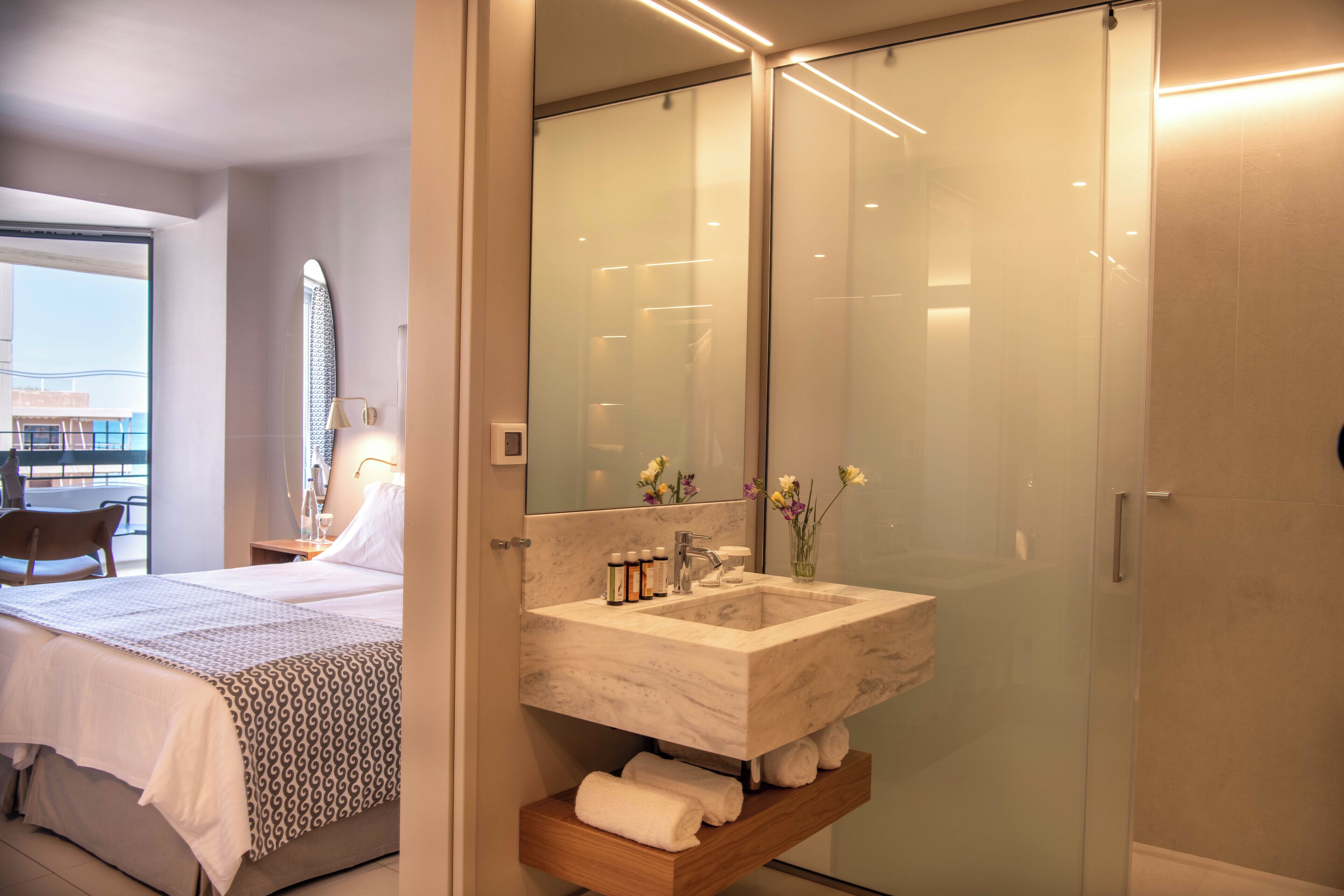 A modern bathroom with a large glass shower enclosure, a marble sink, and a bedside table with toiletries.