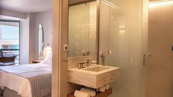 A modern bathroom with a large glass shower enclosure, a marble sink, and a bedside table with toiletries.