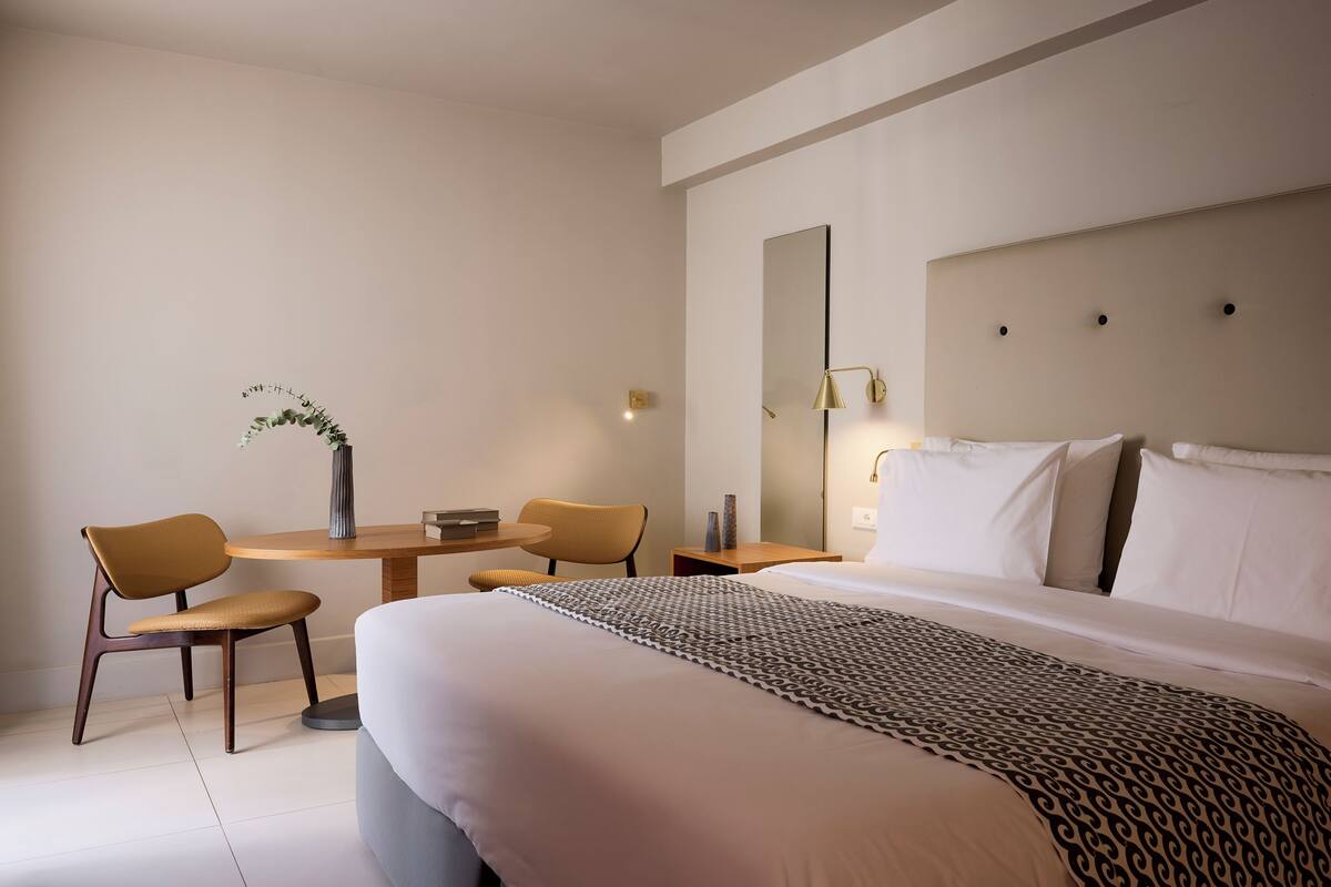 Quarto de hotel moderno com uma cama grande, duas cadeiras, uma mesa pequena e um abajur.