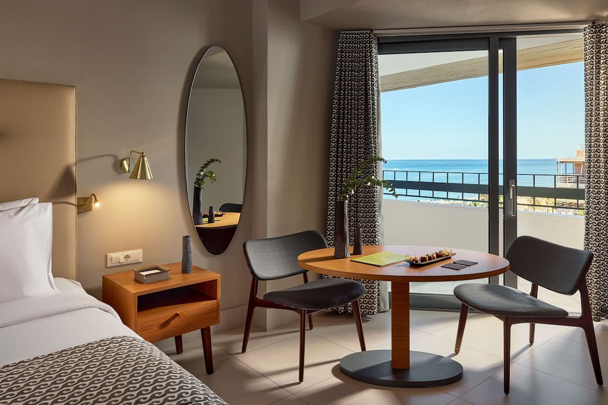 Quarto de hotel com cama, mesa redonda com cadeiras, pequena mesa de cabeceira de madeira, espelho e vista para o mar através de grandes janelas.