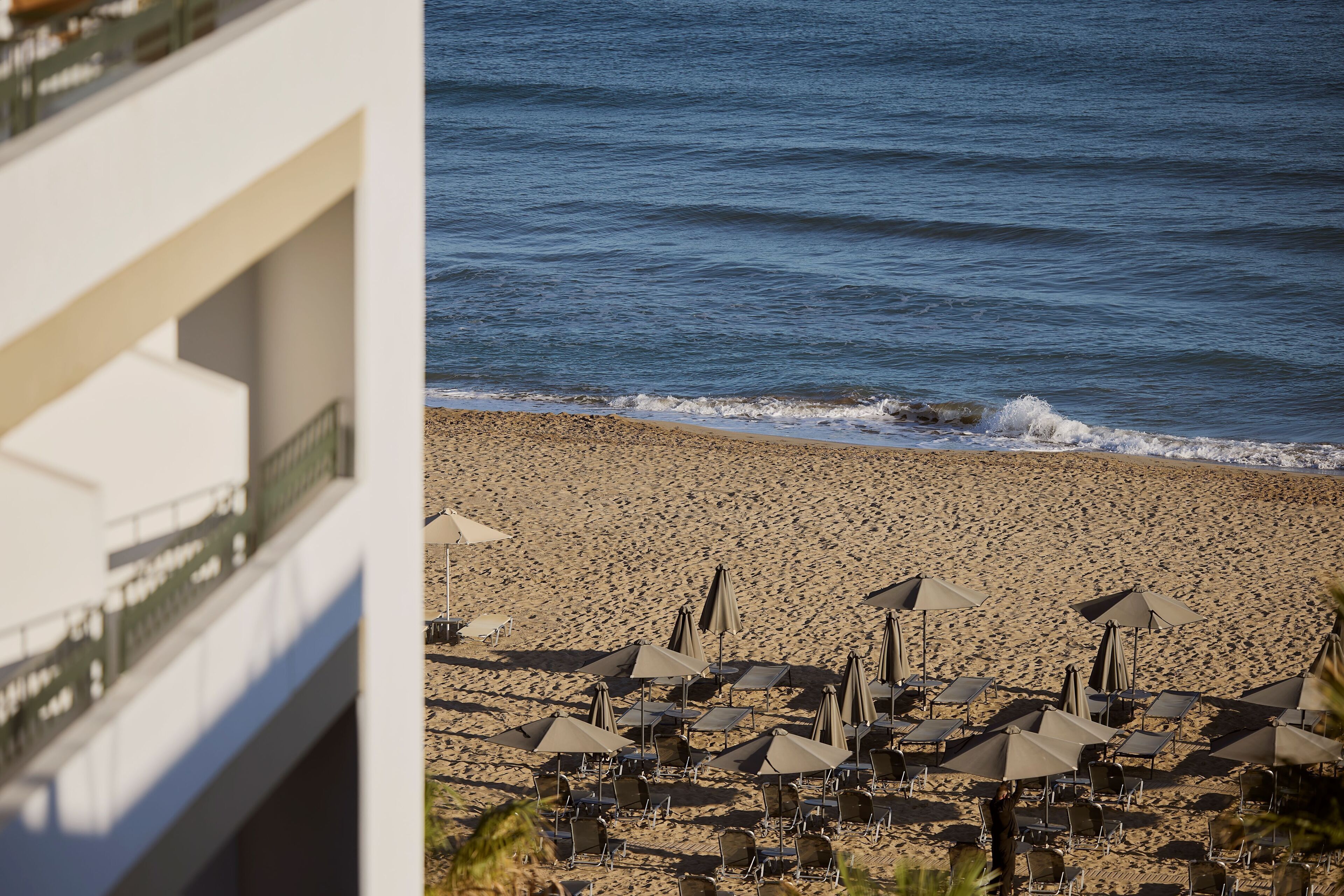 A beach with rows of sun loungers and umbrellas, a clear view of the ocean, and a building in the foreground.