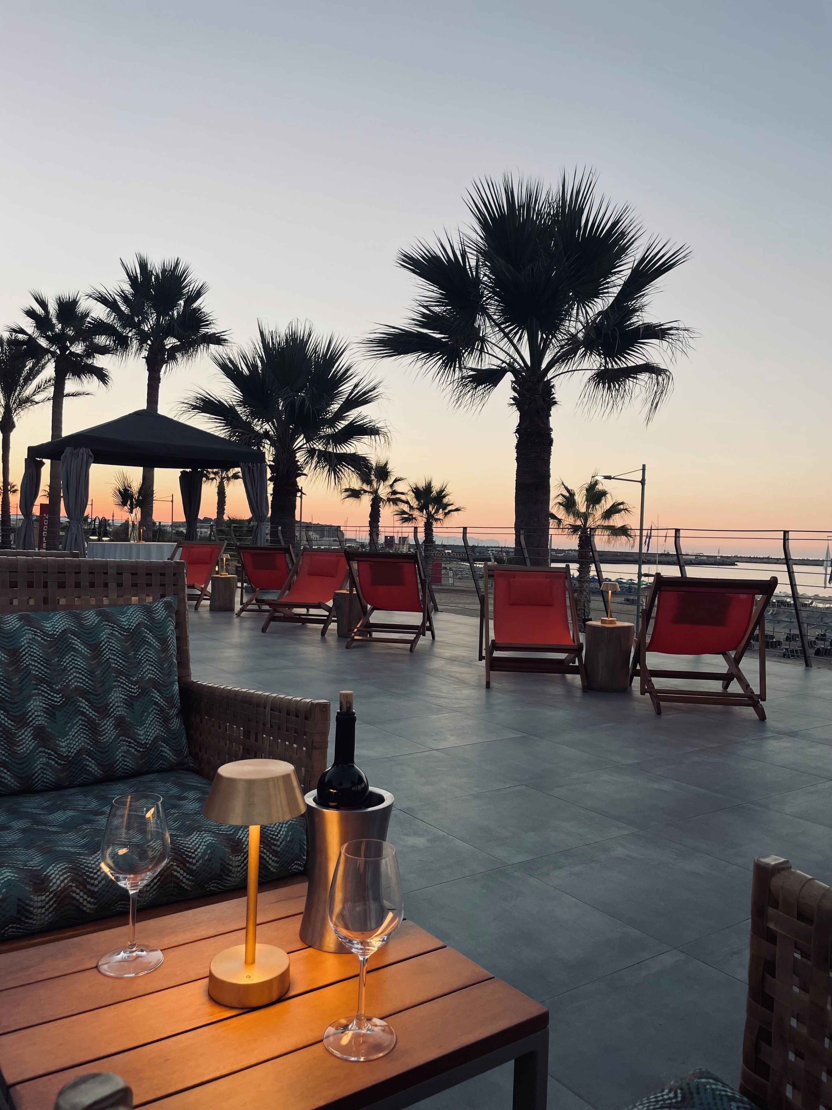 A terrace with red chairs, a table with wine glasses, and a bottle, overlooking a beach with palm trees and a sunset.