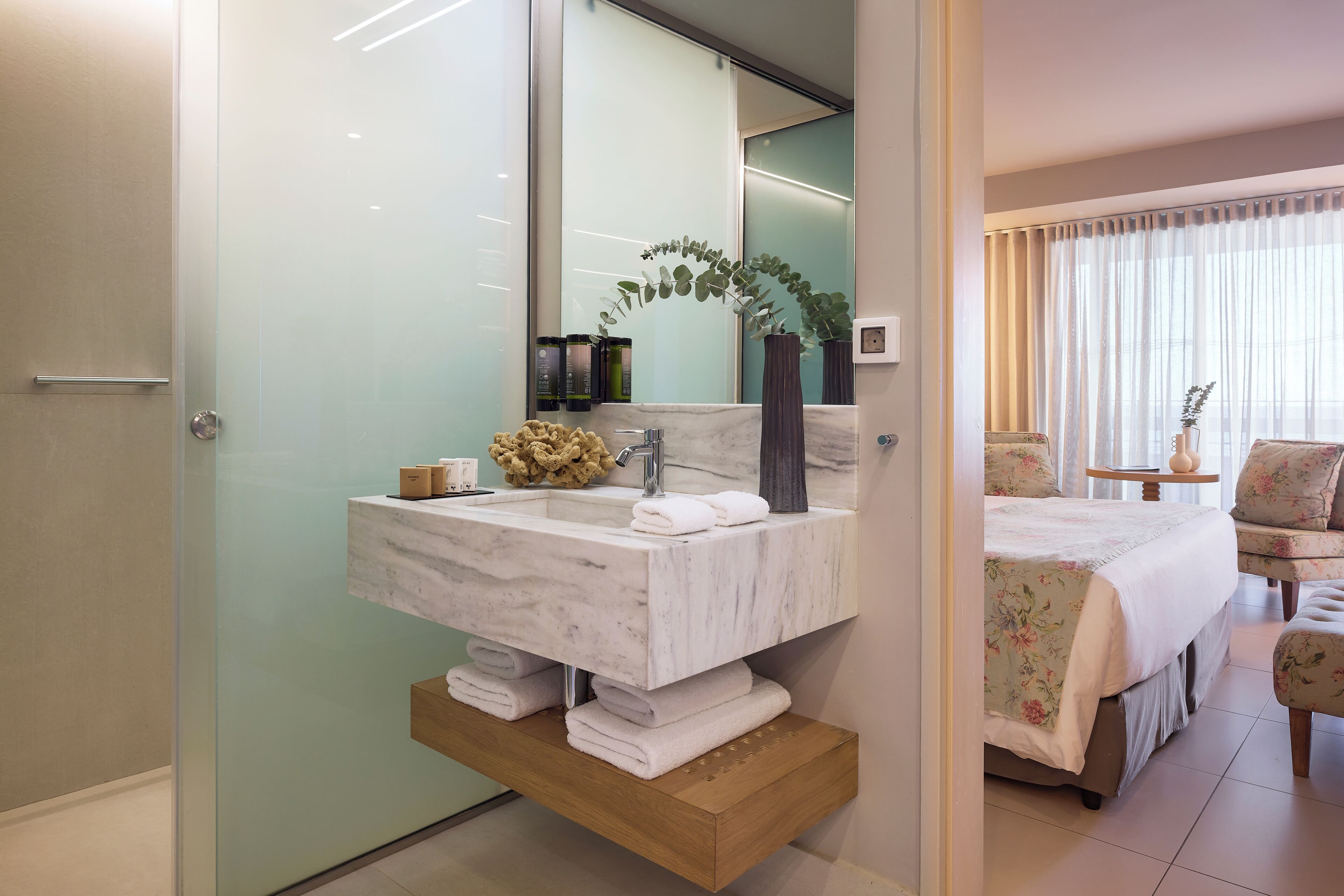 Un baño con lavamanos de mármol, un espejo, toallero y estante de madera.