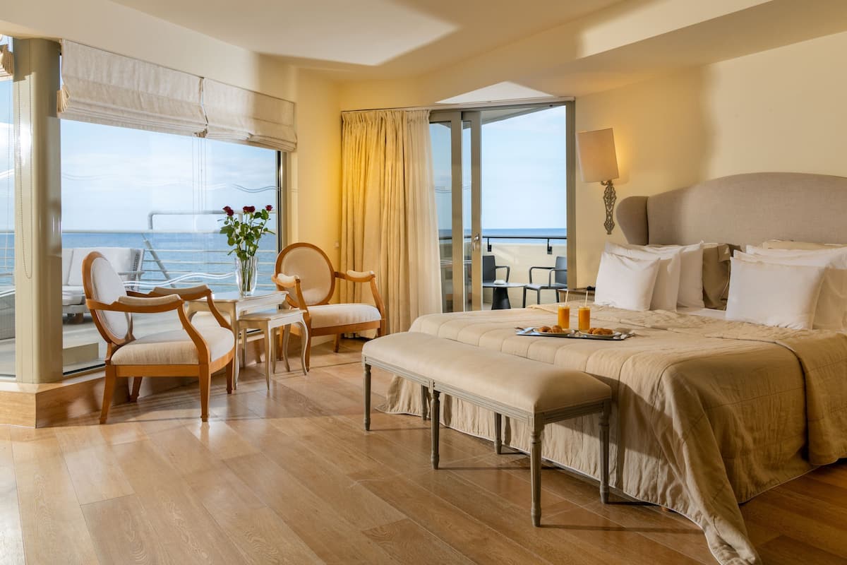 Quarto de hotel com uma cama grande, um espaço de estar com cadeiras, uma mesa e vista para o mar através de uma varanda.